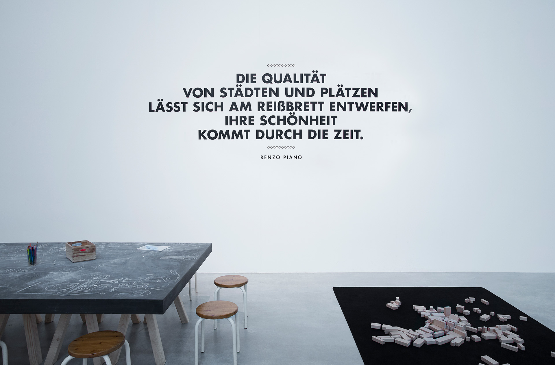 großer Arbeitstisch auf dem viel mit Kreide gekritzelt wurde, daneben schwarze Bodenmatte mit unterschiedlichen Holzblöcken. Dahinter schwarze Schrift auf weißer Wand "Die Qualität von Städten und Plätzen lässt sich am Reißbrett entwerfen, ihre Schönheit kommt durch die Zeit."