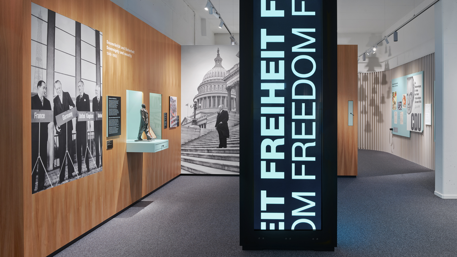 Blick in einen Ausstellungsraum mit hölzernen Wandmodulen und einem Gang rechts. Zentral steht ein schmales, hohes, schwarzes Ausstellungsmöbel mit zwei übereinander angeordneten Displays, auf denen ‚Freiheit – Freedom‘ steht.
