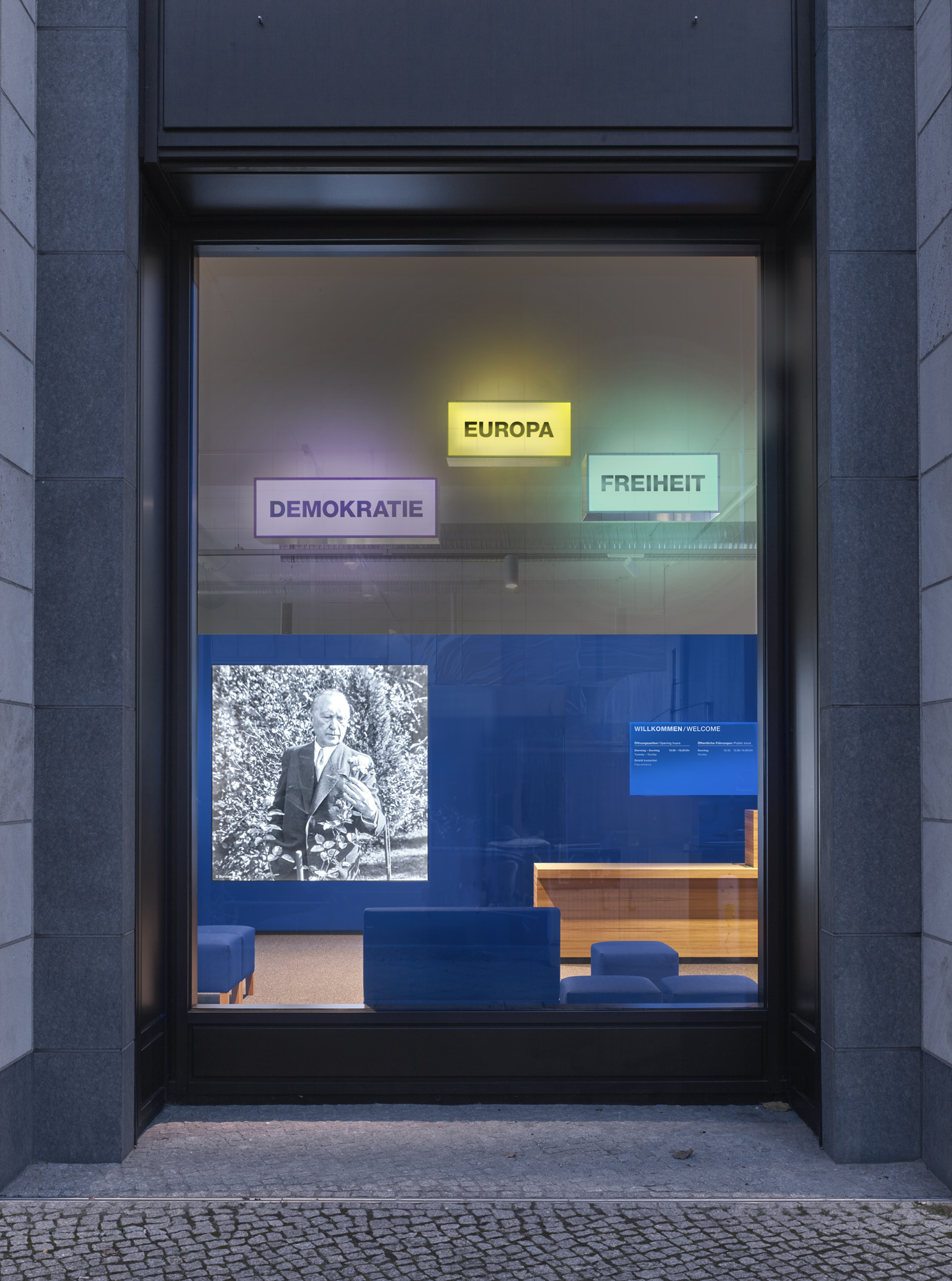 Schaufenster von außen mit 3 Leuchtboxen "Demokratie", "Europa" und "Freiheit". Dahinter eine blaue Wand mit einem großen Display auf dem Konrad-Adenauer in schwarz-weiß zu sehen ist.