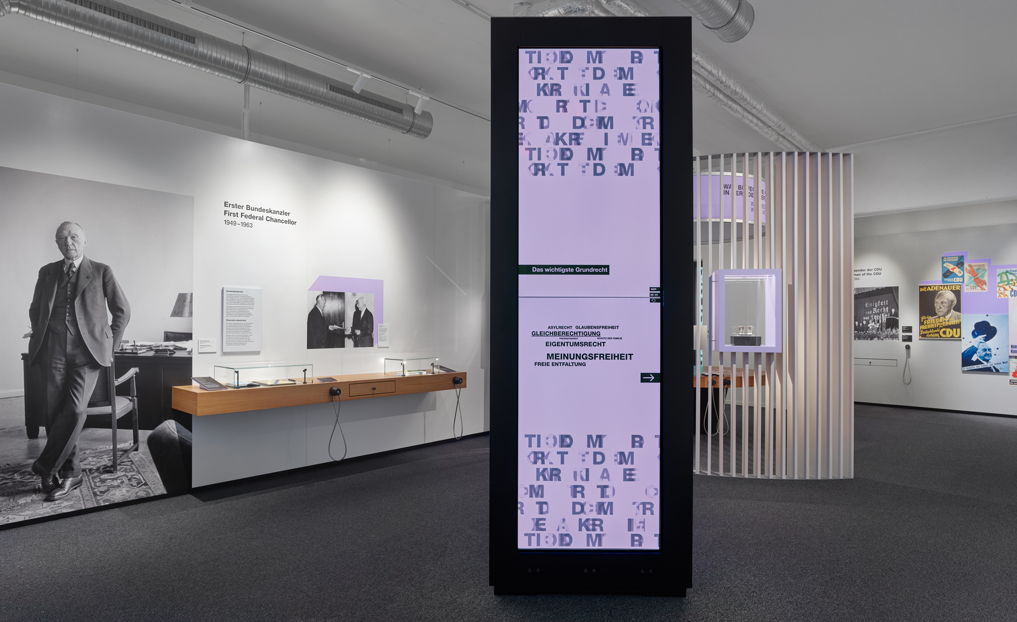 Blick in einen Ausstellungsraum. Zentral steht ein schmales, hohes, schwarzes Ausstellungsmöbel mit zwei übereinander angeordneten Displays, auf denen verschiedene demokratische Begriffe zu sehen sind.