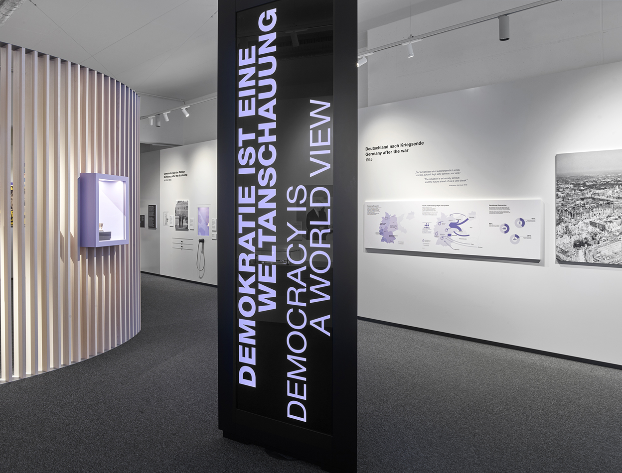 Blick auf ein schmales, hohes, schwarzes Ausstellungsmöbel mit zwei übereinander angeordneten Displays, auf dem in violetter Schrift "Demokratie ist eine Weltanschauung" steht.