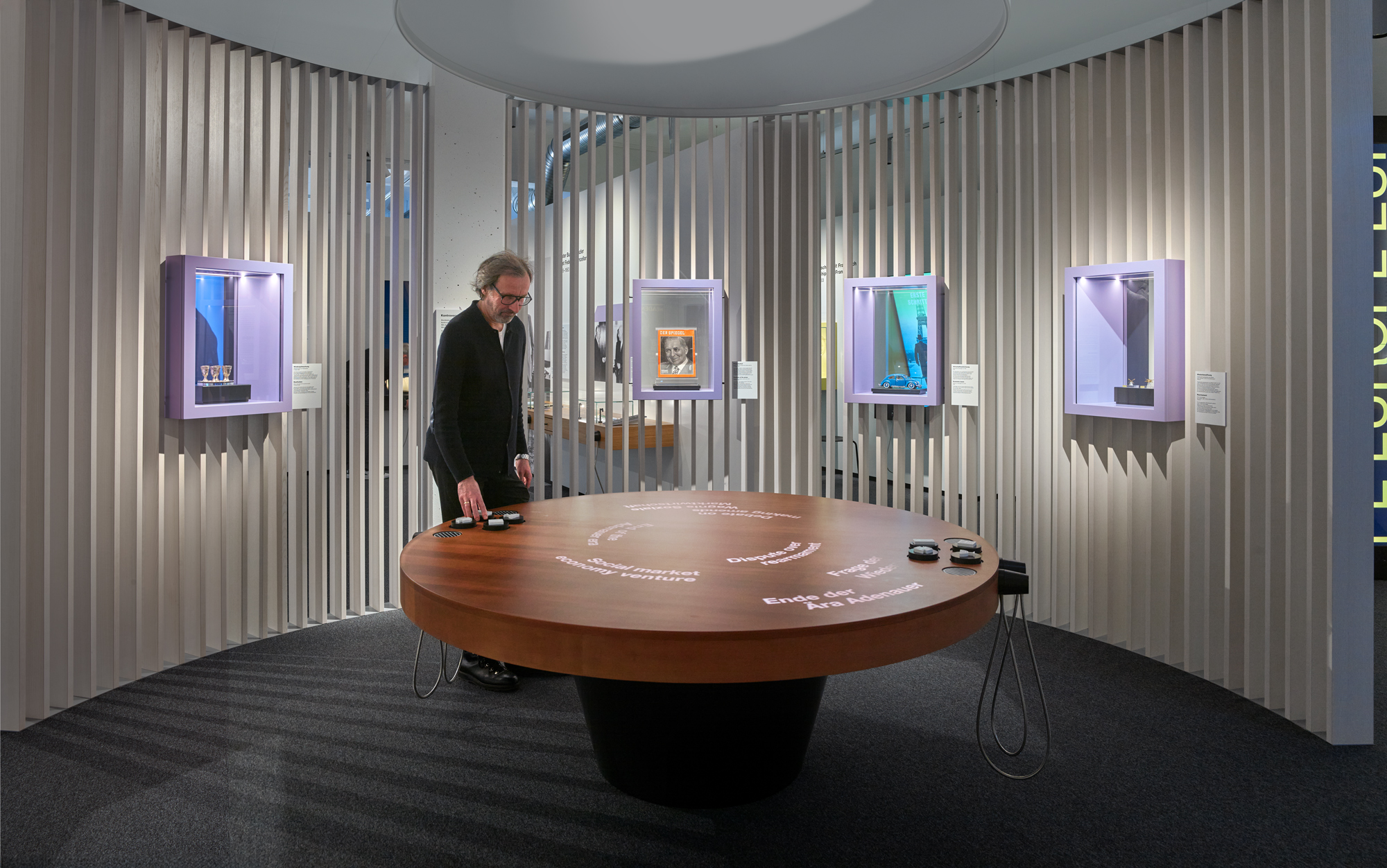 Großer runder hölzener Tisch mit einer Projektion darauf, mehreren Einhandhörern und verschiedenen Tokens darauf. Eine männliche Person steht dahinter und interagiert mit dem Tisch und den Tokens.