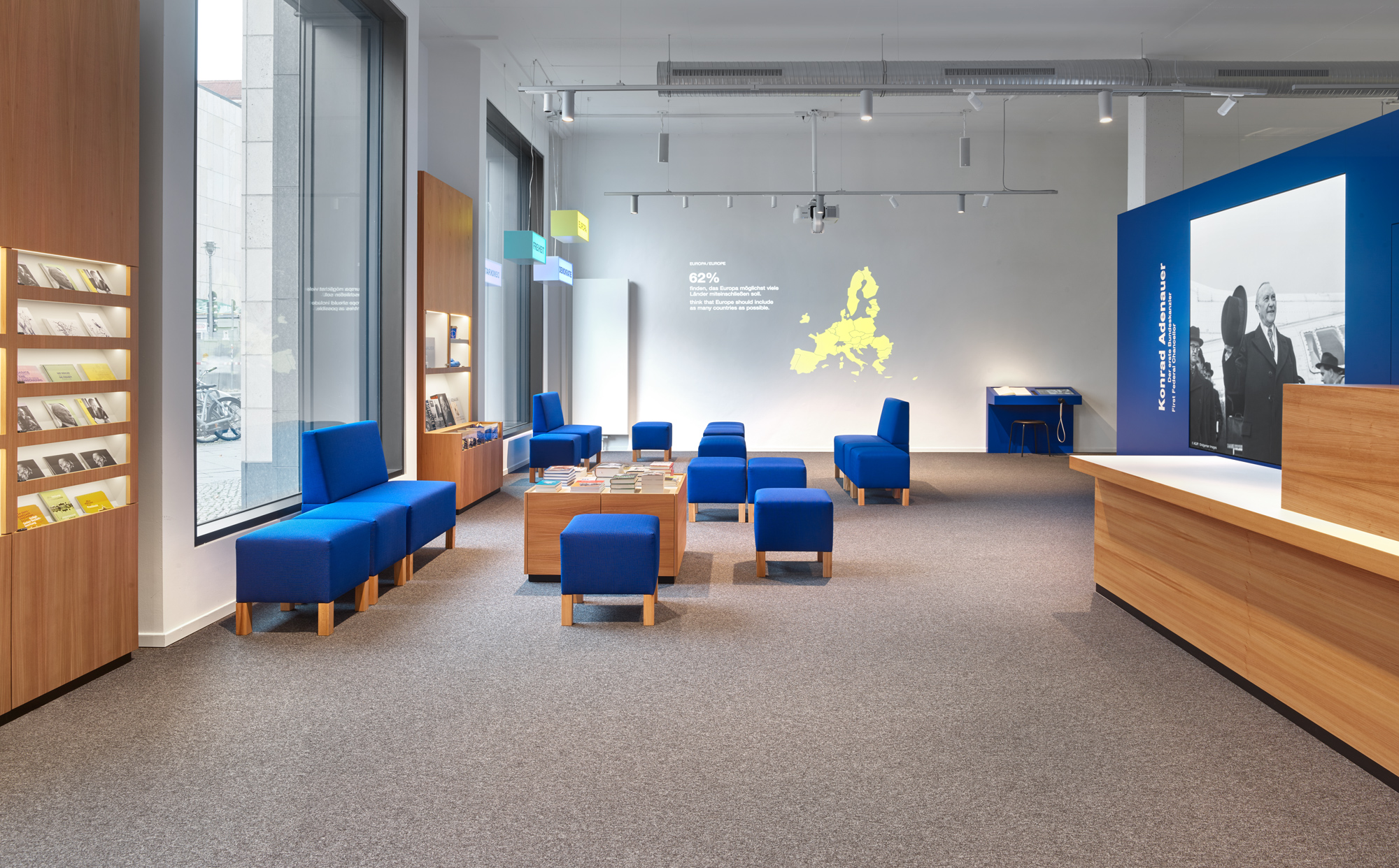 Forum mit rechteckigen, blauen Sitzmöbeln unterschiedlicher Größe. Rechts ein Empfangstresen, dahinter ein großes Bild von Konrad Adenauer. An der zentralen Wand eine Projektion mit Statistiken und einer Europakarte.