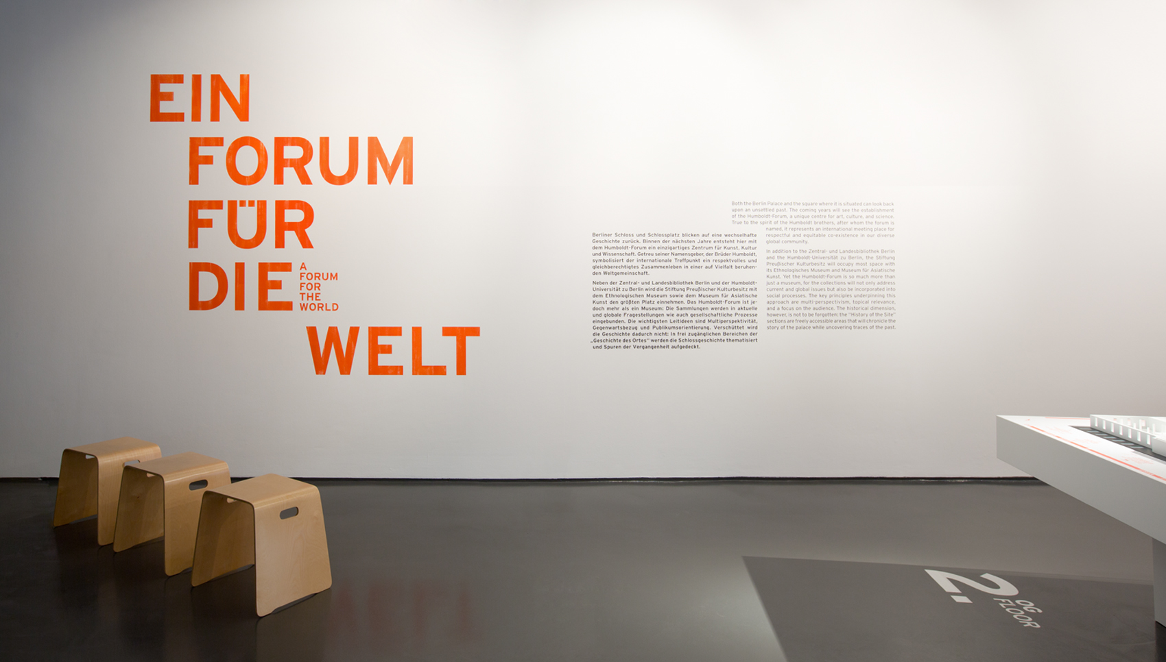 roter Wandtext "Ein Forum für die Welt" auf weißer Wand. Davor Holzhocker