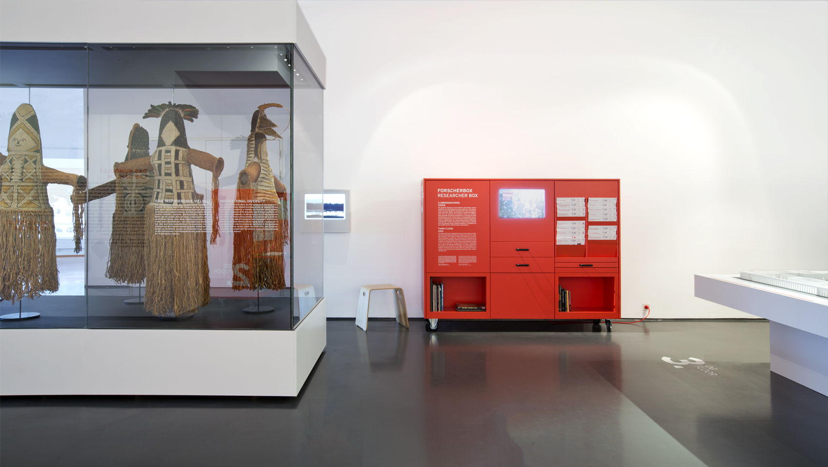 Mobiles rotes Ausstellungsmöbel mit verschiedenen Auszügen, Texten, Fächern für Bücher und einem Monitor. Davor große Schrankvitrine mit traditioneller Bekleidung