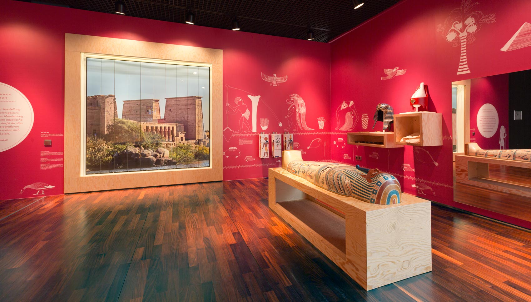 rote Ausstellungswand mit weißen Grafiken darauf. großes Ausstellungsmöbel mit Sarkophag davor