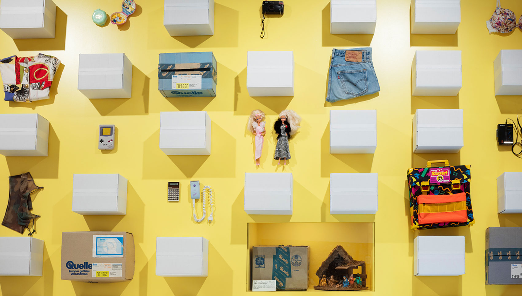 gelbe Wand mit verschiedenen Exponaten. Darunter ein Scout-Rucksack, Barbie-Puppen, ein Gamboy, ein BH etc.