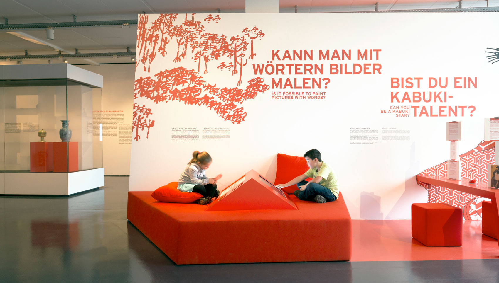 rote Sitzinsel mit Medienstation auf der zwei Kinder sitzen und damit interagieren