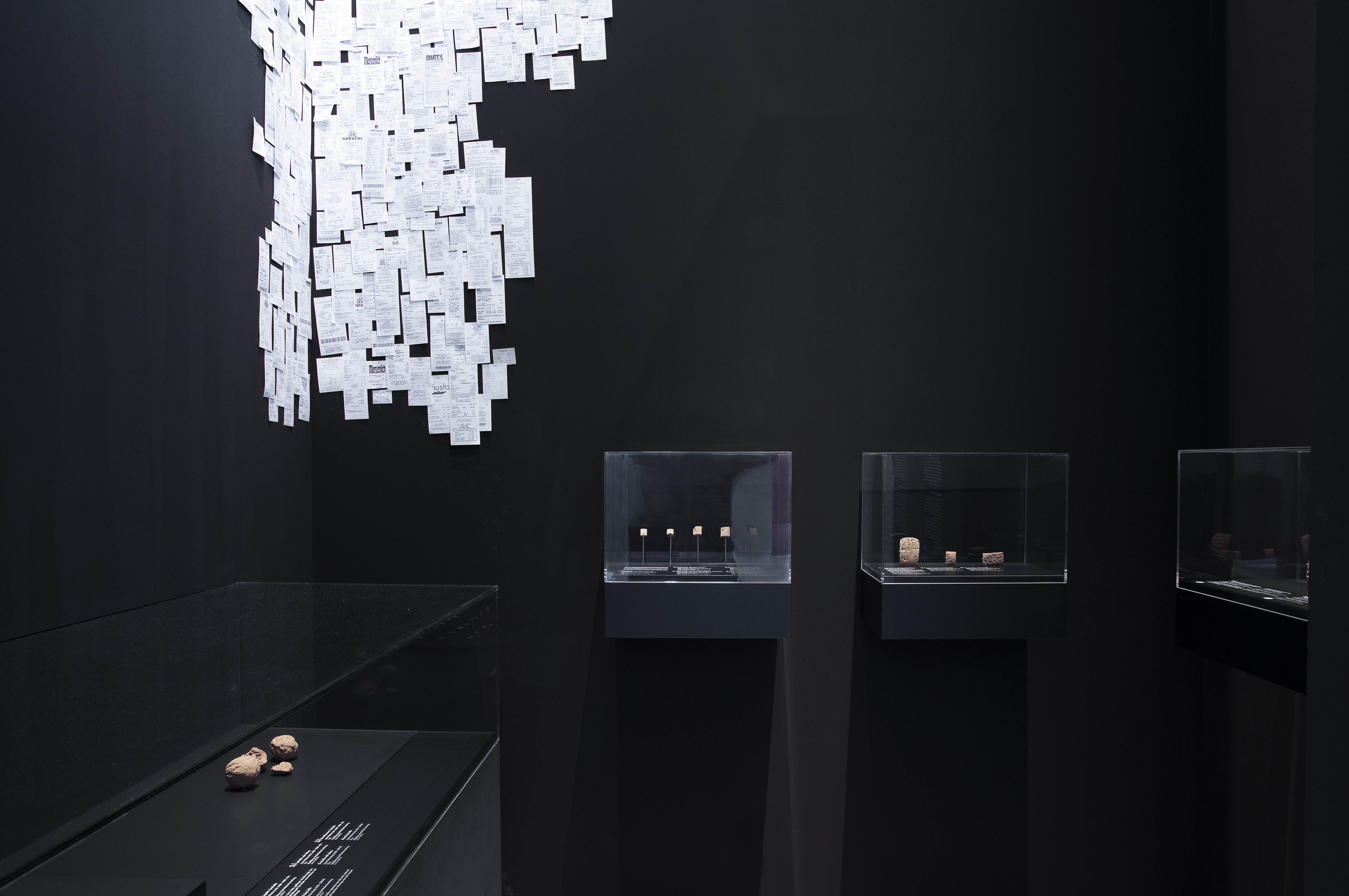 Schwarzer Raum mit schwarzen Aufsatzvitrinen. In Raumecke Installation aus weißen Einkaufsquittungen