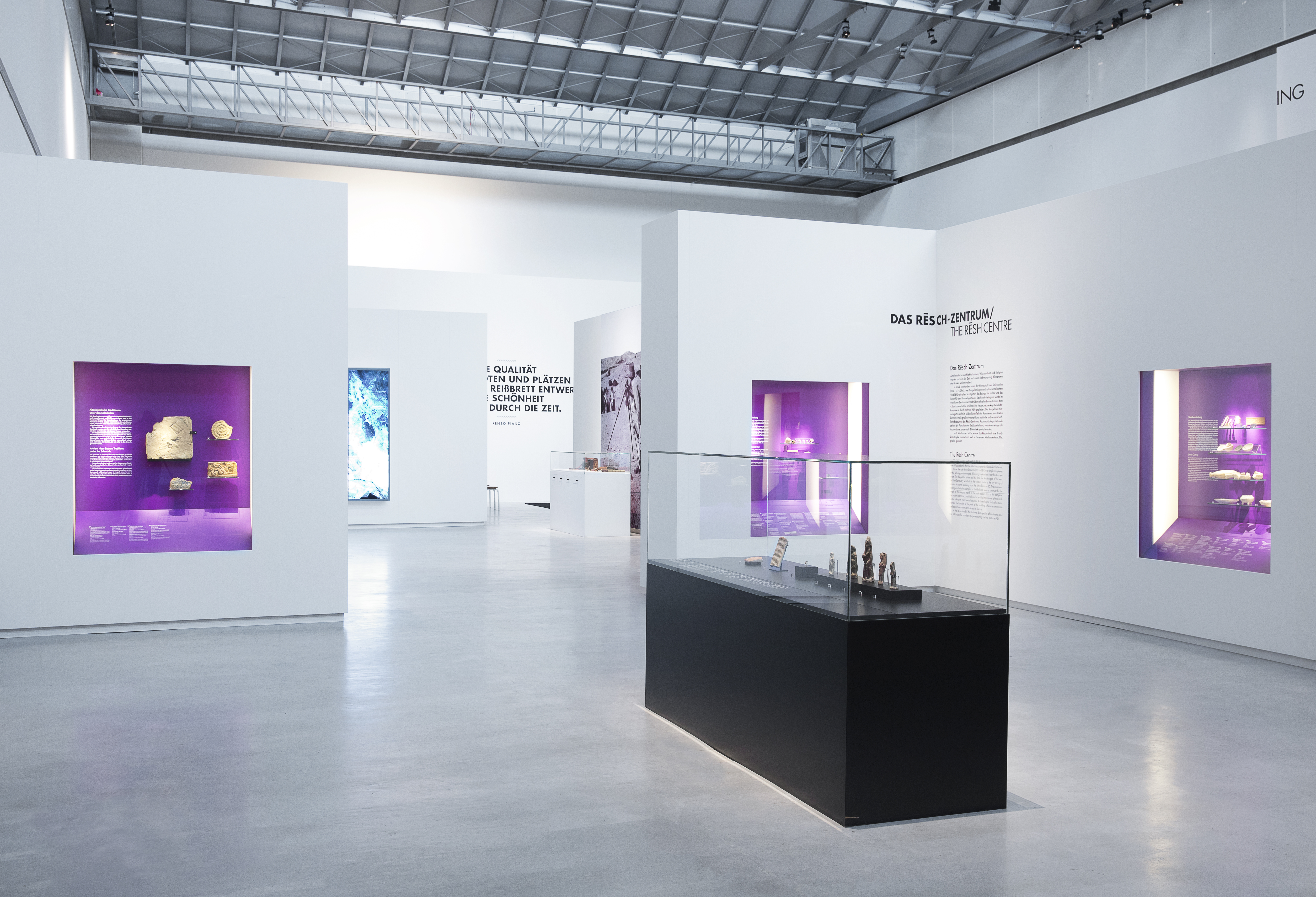 weiße Ausstellungswände mit Wandvitrinen, die sich durch violette Rückwand abheben. Im Raum Aufsatzvitrinen Möbel in schwarz.