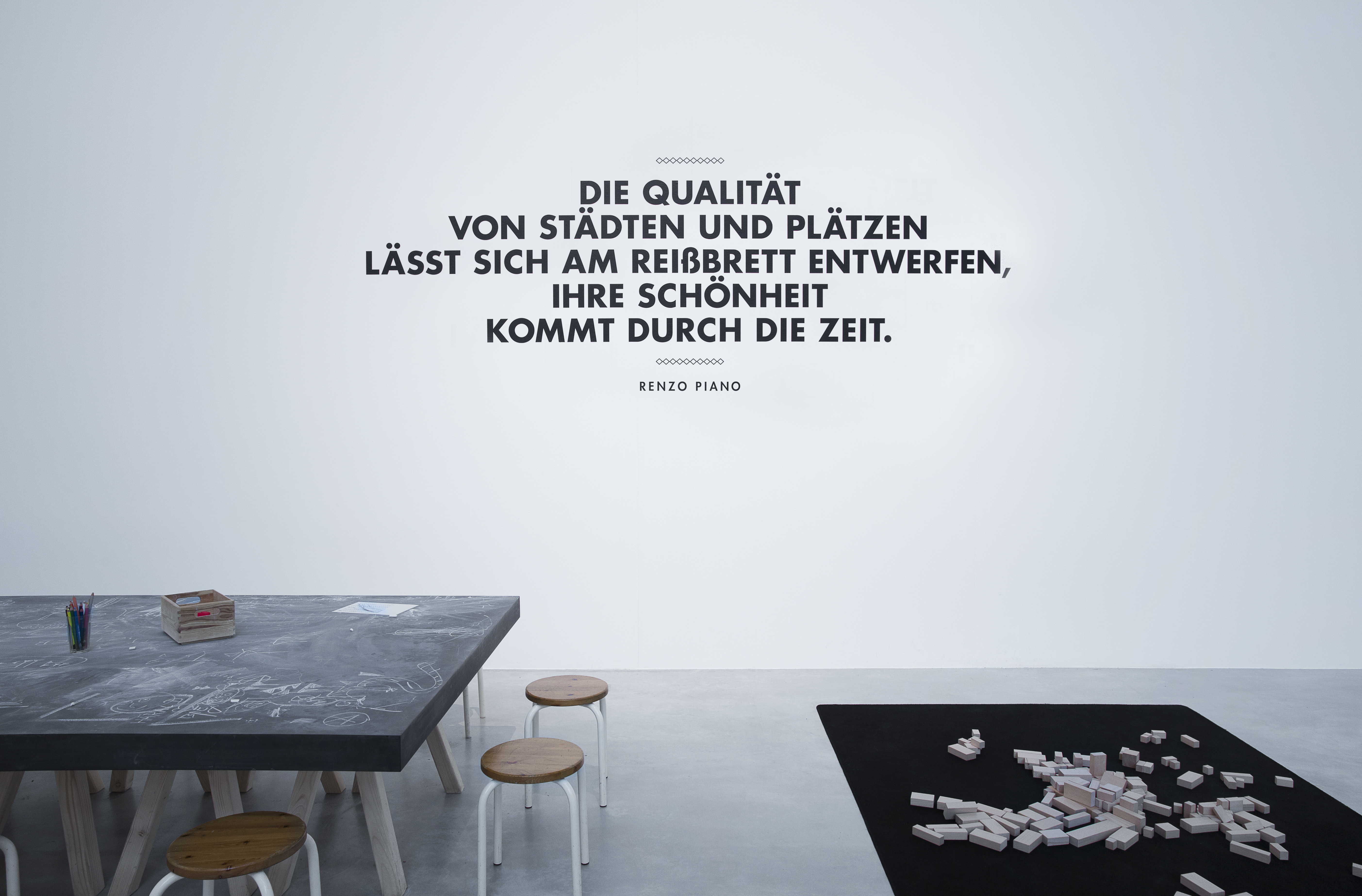 großer Arbeitstisch auf dem viel mit Kreide gekritzelt wurde, daneben schwarze Bodenmatte mit unterschiedlichen Holzblöcken. Dahinter schwarze Schrift auf weißer Wand "Die Qualität von Städten und Plätzen lässt sich am Reißbrett entwerfen, ihre Schönheit kommt durch die Zeit."