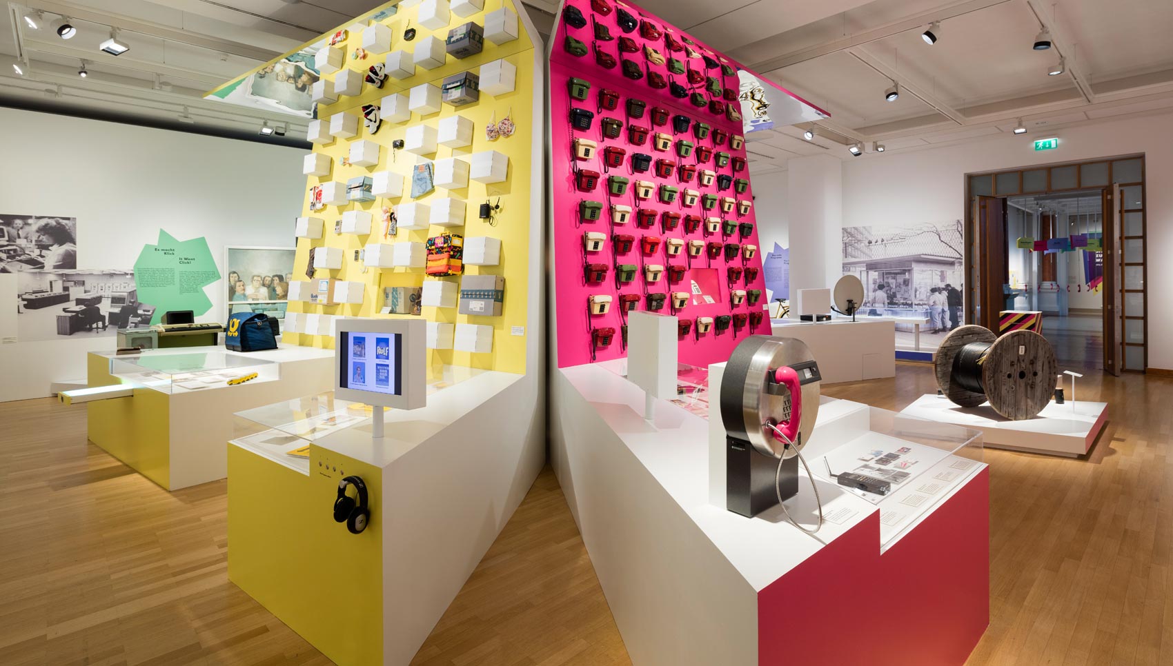 Blick auf gelbe Wand mit gemischten Exponaten und pinke Wand mit Telefonen
