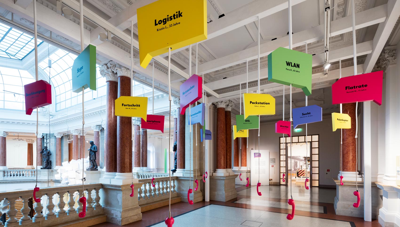 Rauminstallation mit eckigen Sprechblasen an denen Magentafarbene Schnurtelefone runterhängen