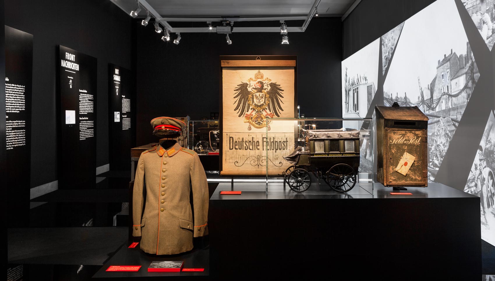 Ausstellungsraum mit schwarzen Wänden. Links Schwarze Paneele mit weißem Text. Zentral Tischpodest mit Exponaten (zum Beispiel Uniform) und Vitrinen. Rechts große Projektion