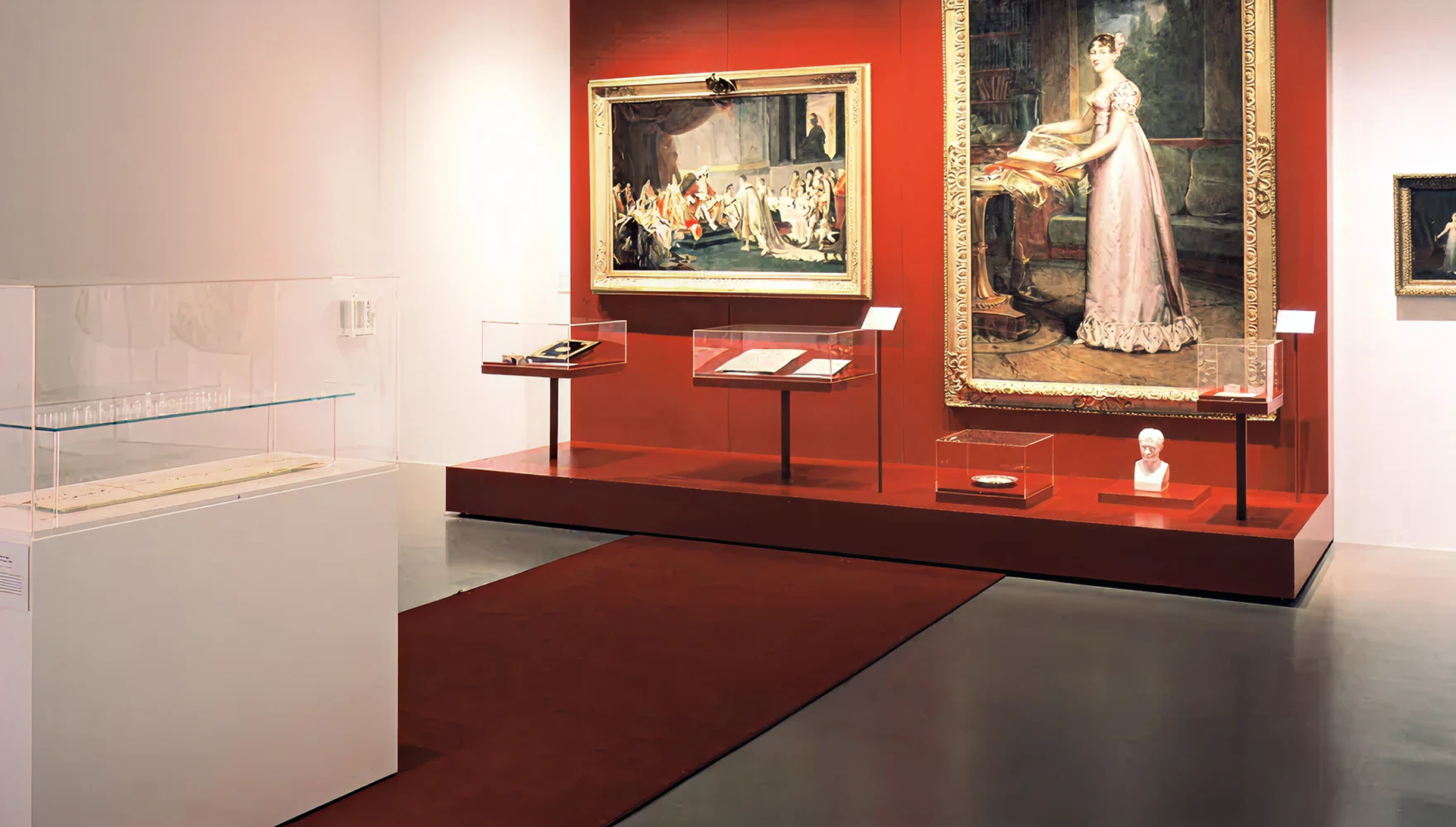 Blick auf verschiedene Vitrinen und Exponate. Dahinter rote Wand mit zwei sehr großen Gemälden in goldenen Rahmen