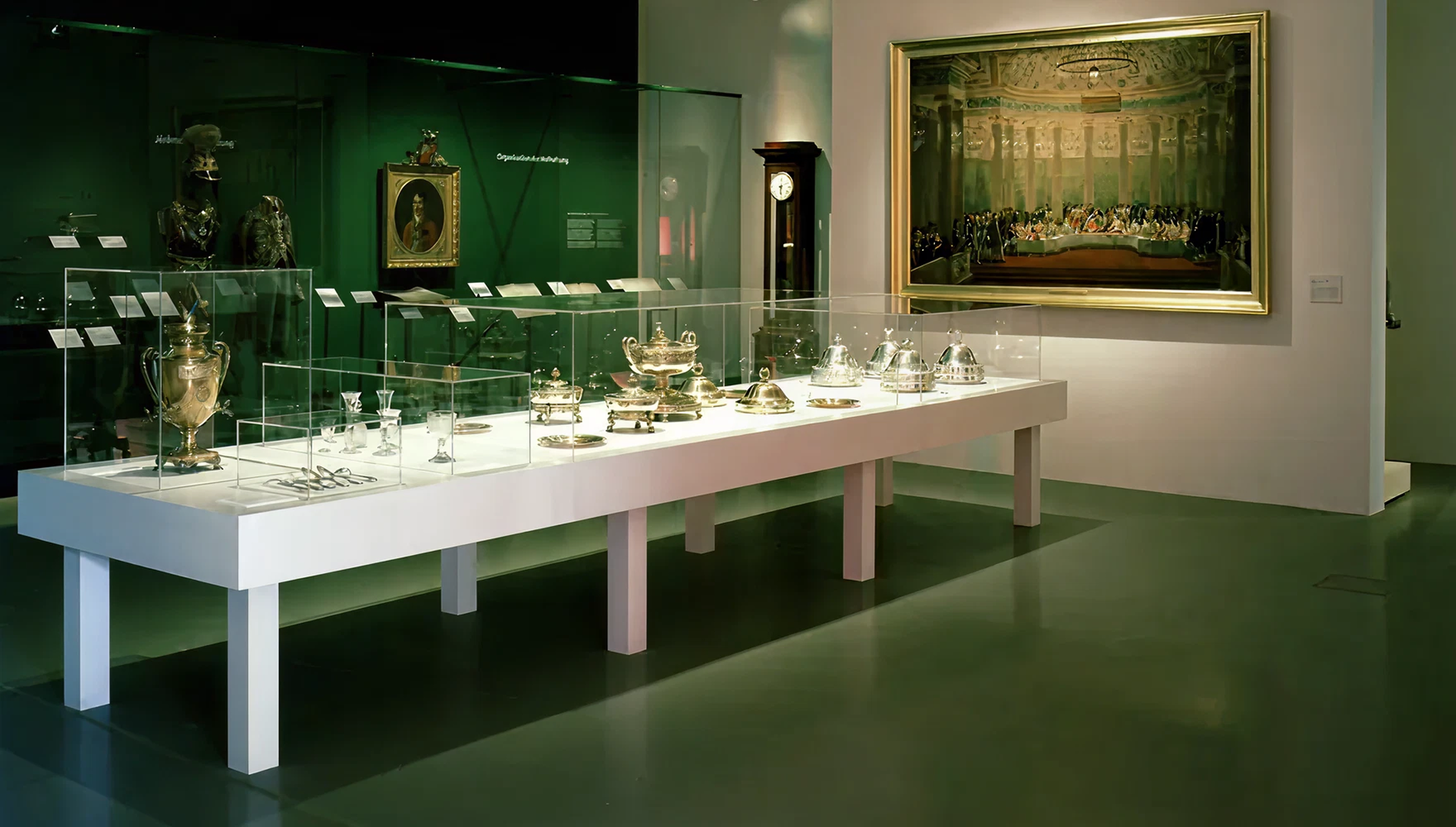 Blick in einen Raum mit dunkelgrünem Boden und dunkelgrüner Wand. Zentral ein großer Ausstellungstisch mit verschiedenen Aufsatzvitrinen und unterschiedlichen Exponaten mit dem Thema Silbergeschirr.