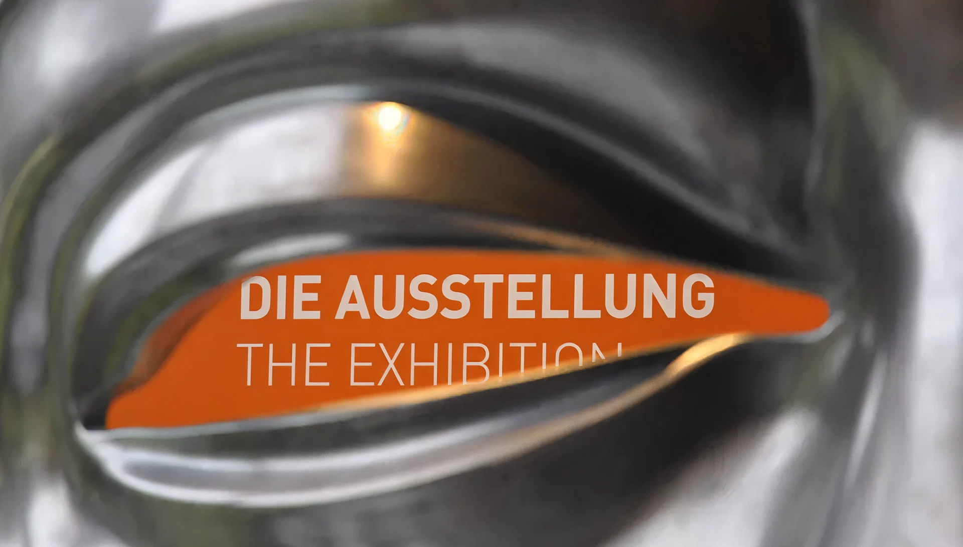 Blick durch Stahl Auge auf weißen Text vor orangener Wand "Die Ausstellung"