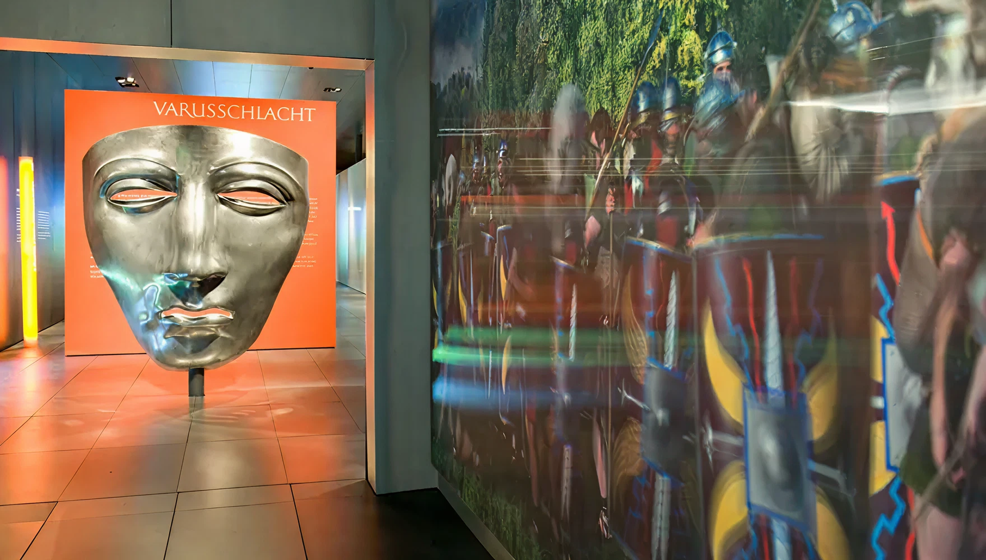 überdimensionale Stahlmaske vor orangener Wand mit dem Titel "Varusschlacht"