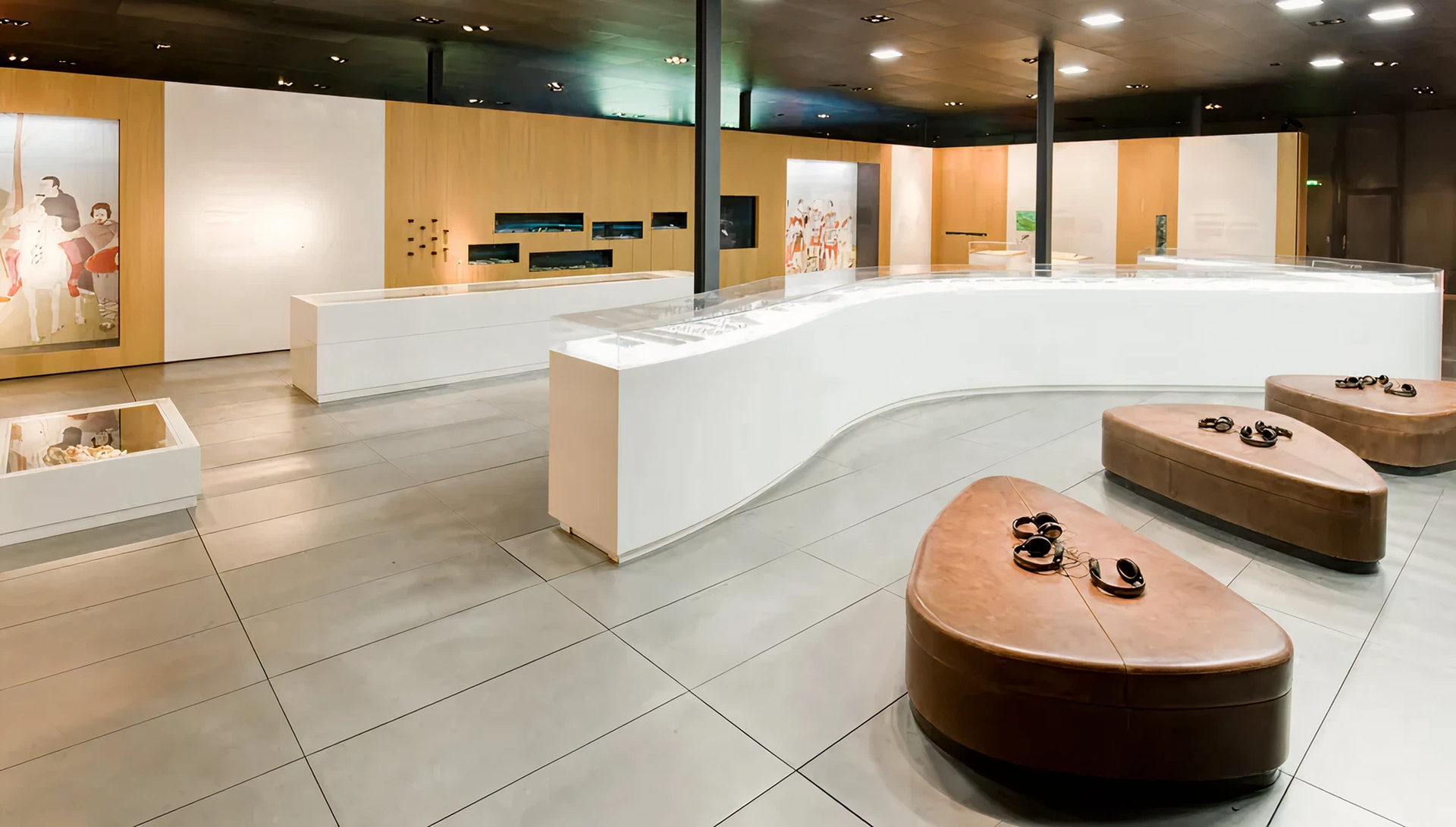 Blick in Ausstellungsraum auf verschiedene braune, lederne Sitzmöglichkeiten mit Kopfhörern darauf, weiße Tischpodeste mit Vitrinen und hölzerne Ausstellungswände.