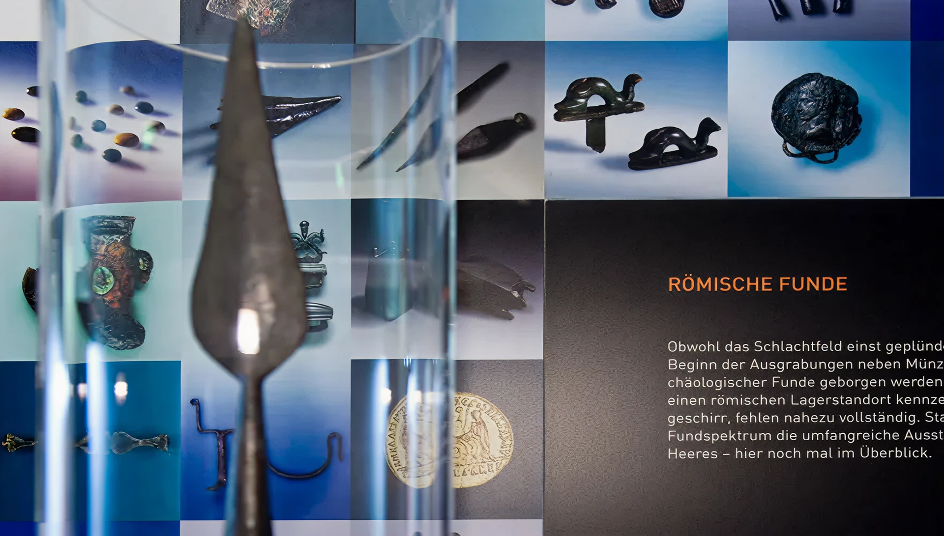 Blick auf Fotowand mit dem Thema "Römische Funde". Davor runde Aufsatzvitrine mit einer eisernen Speerspitze.