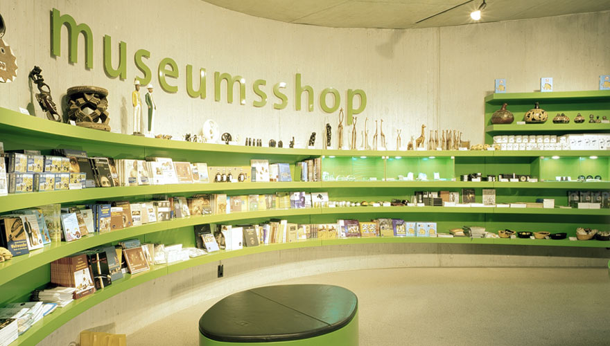 Eine große, grüne Regalwand, in der verschiedene Souvenirs zu finden sind. Darüber an der Wand steht „Museumsshop”.