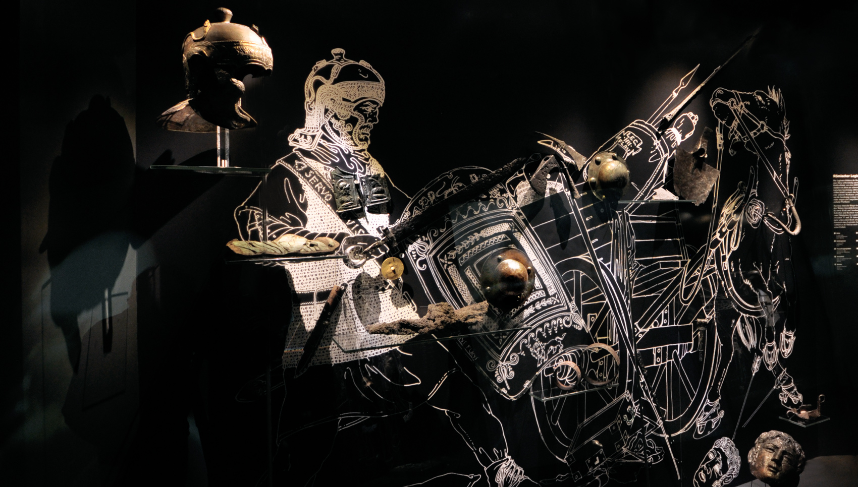 weiße Umzeichnung eines römischen Kriegers auf schwarzer Wand mit Exponaten davor, die Teile seiner Ausrüstung darstellen