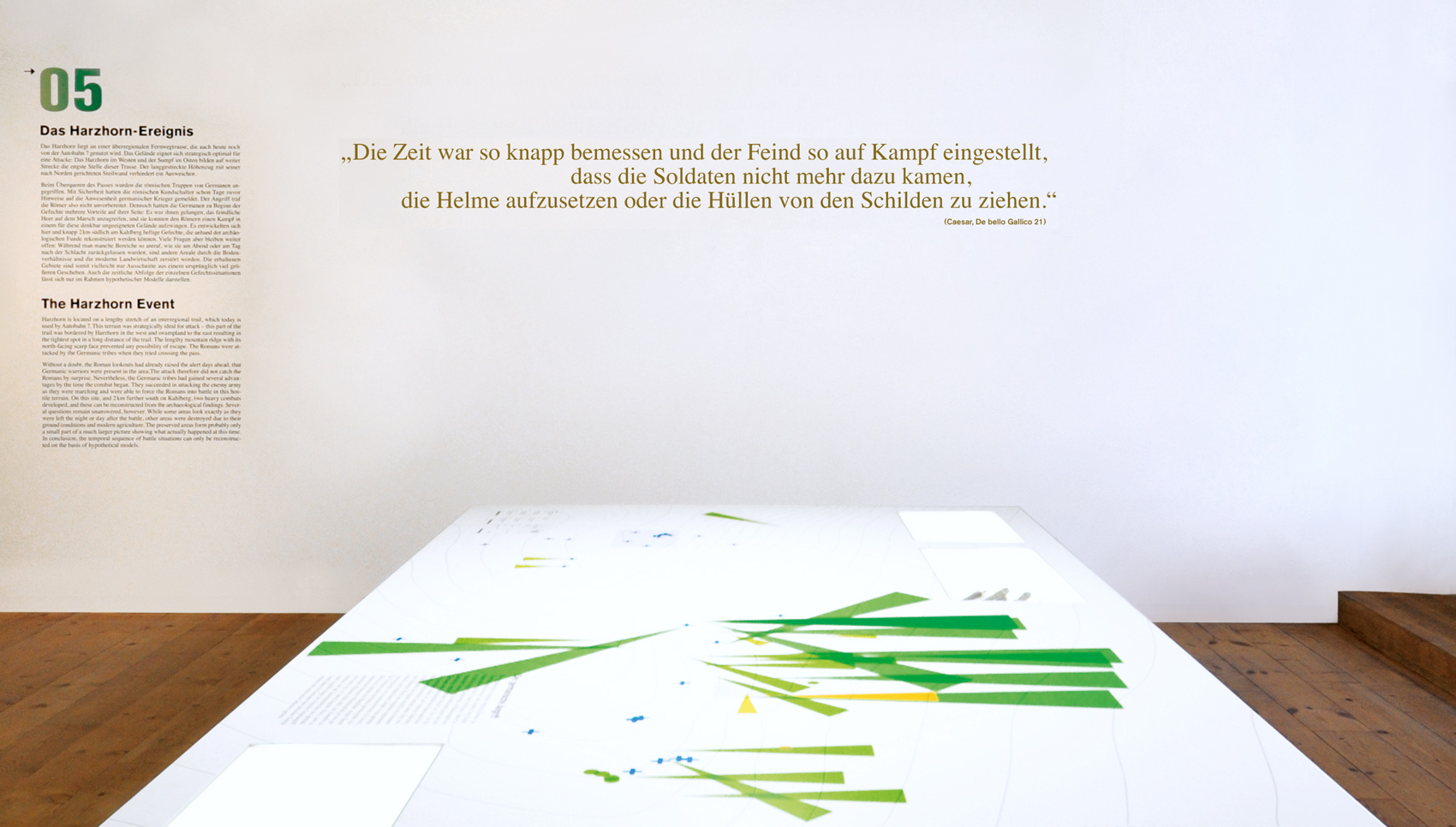 weißer Tisch mit topografischer Karte und grünen Pfeilen vor weißer Wand mit Text "Die Zeit war so knapp bemessen und der Feind so auf Kampf eingestellt, dass die Soldaten nicht mehr dazu kamen, die Helme aufzusetzen oder die Hüllen von den Schilden zu ziehen."