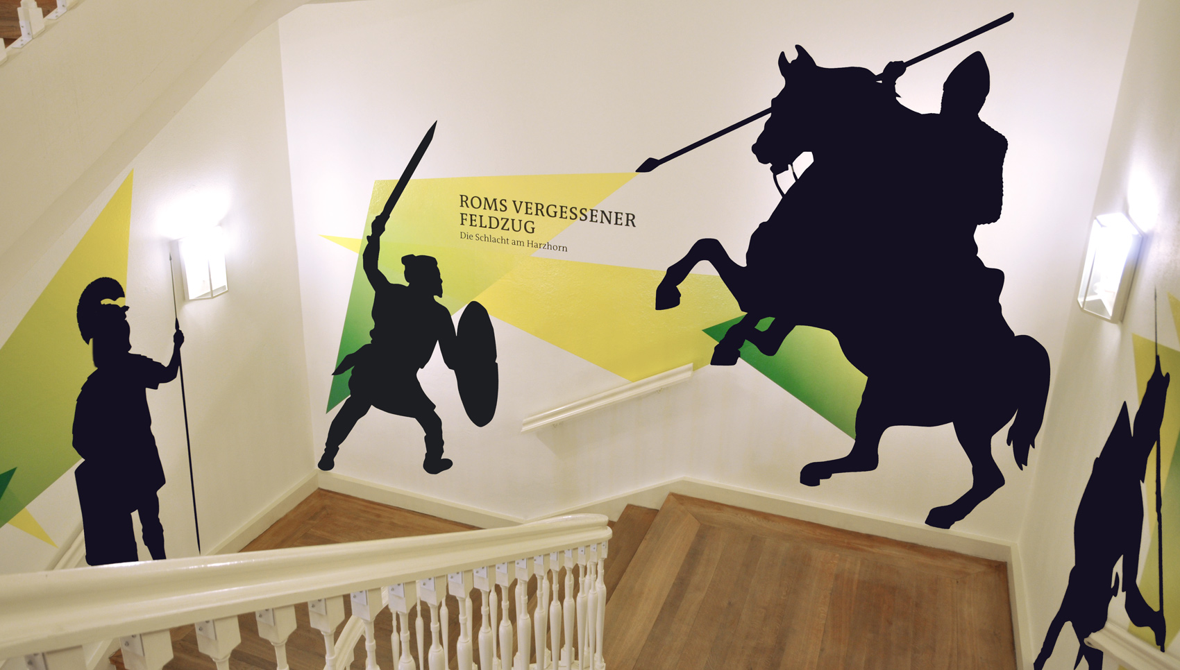 Treppe von oben nach unten mit Blick auf Wandgrafik "Roms vergessener Feldzug". Schwarze Silhouette von Kämpfern