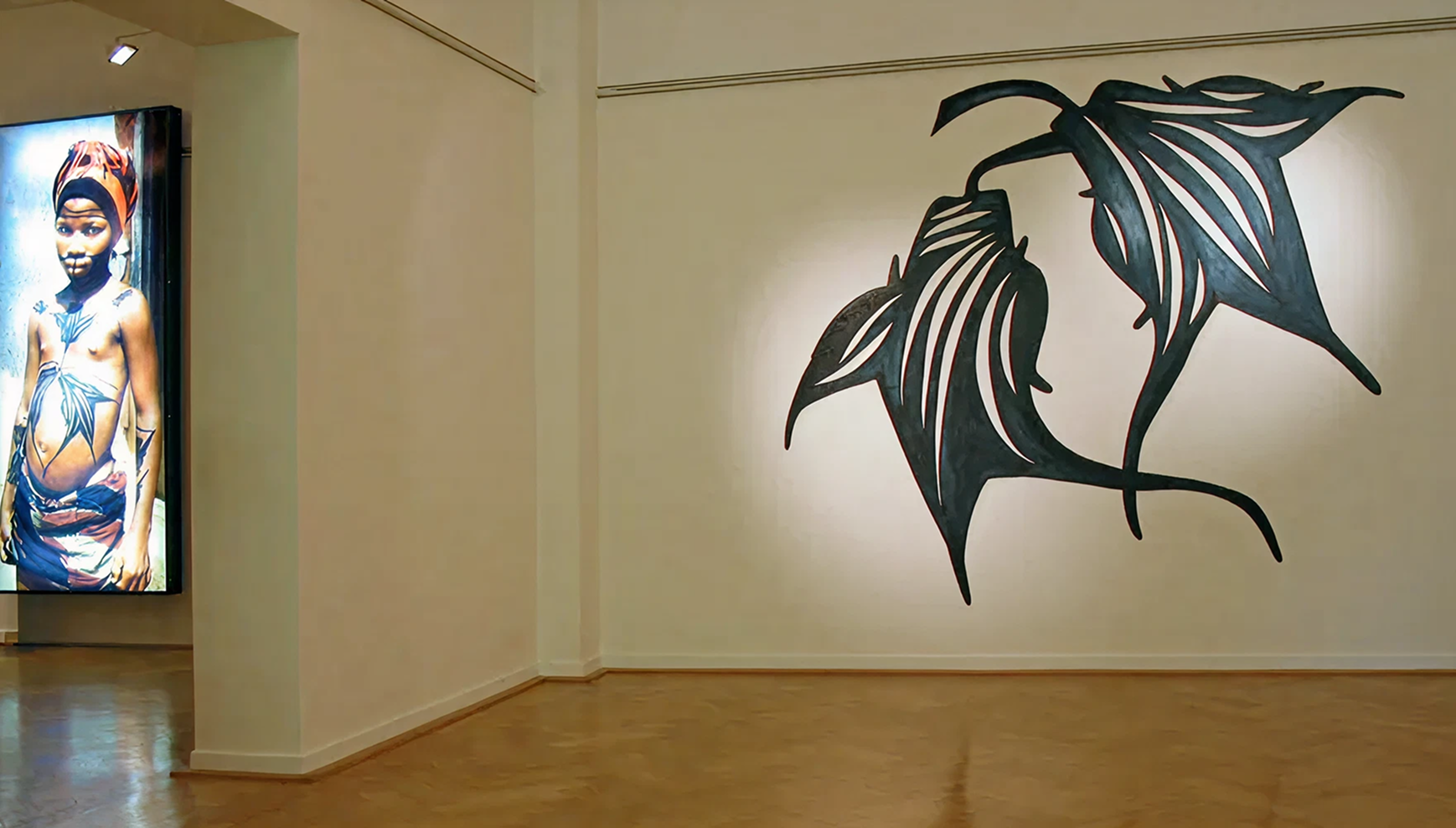 Wandgrafik eines überdimensionalen Tattoos neben einer leuchtenden Fotobox. Zu sehen ist eine indigene Person, die das auf der Wandgrafik abgebildete Tattoo auf dem Bauch trägt.