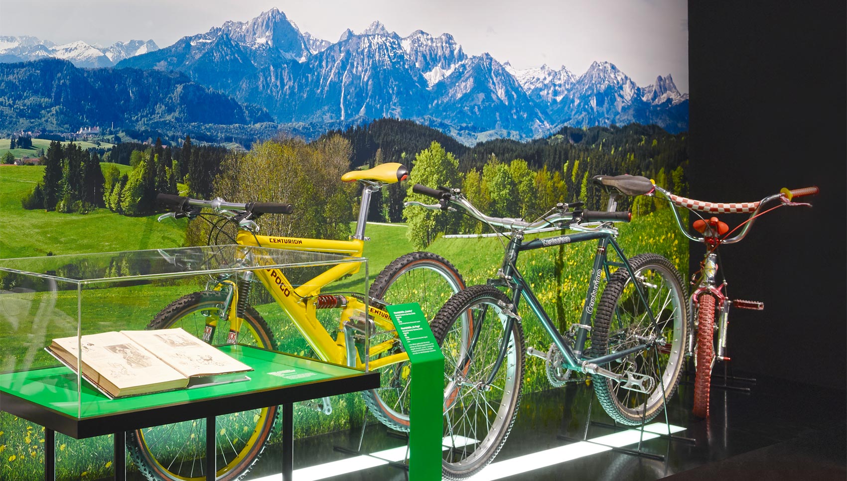 Ausstellungsausschnitt mit Fahrrad Exponaten und Vitrine vor einer Grafikwand