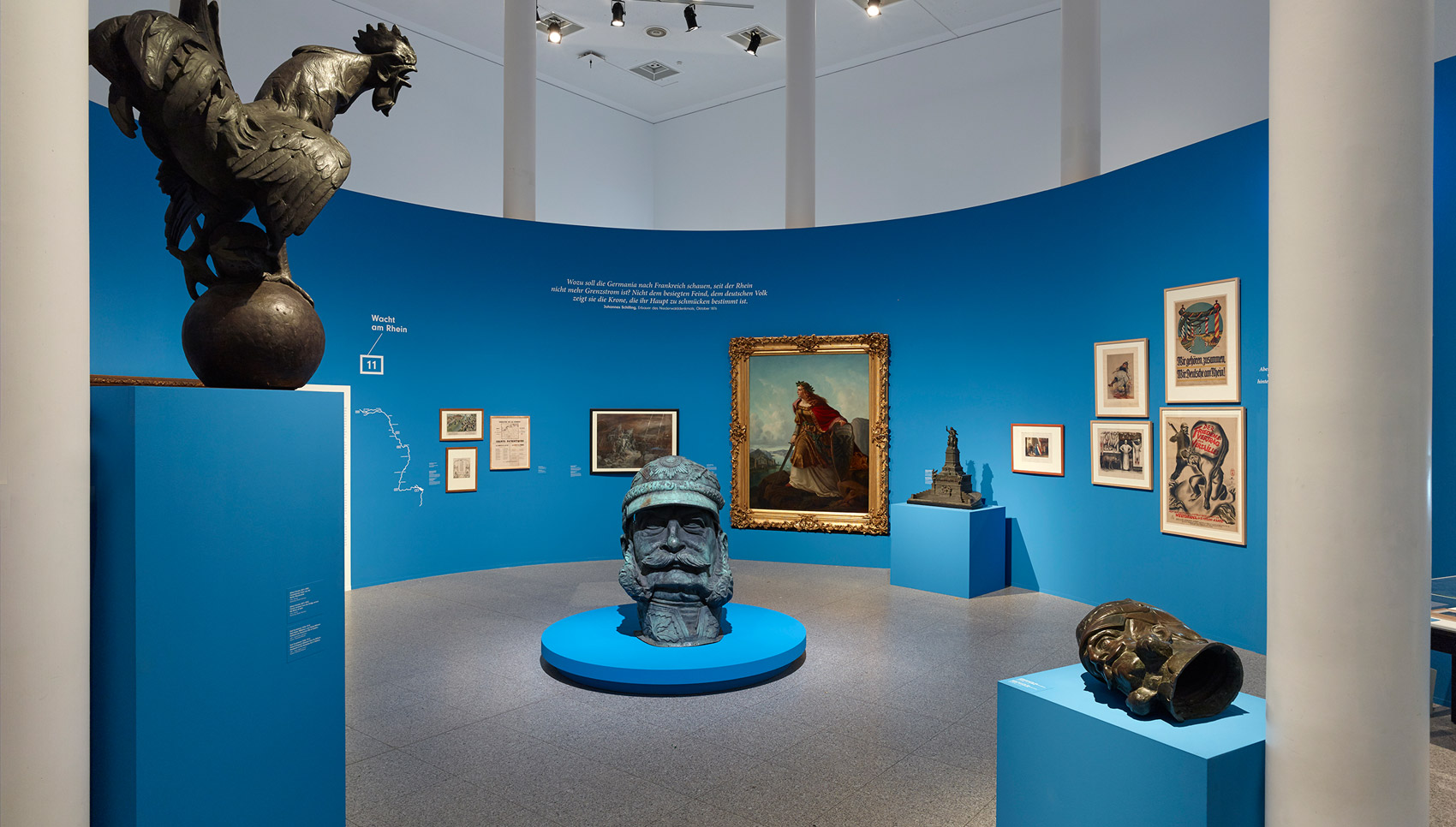 In einem blauen Ausstellungsraum hängen Gemälde und stehen Exponate. In der Mitte steht eine große Kopfskulptur.