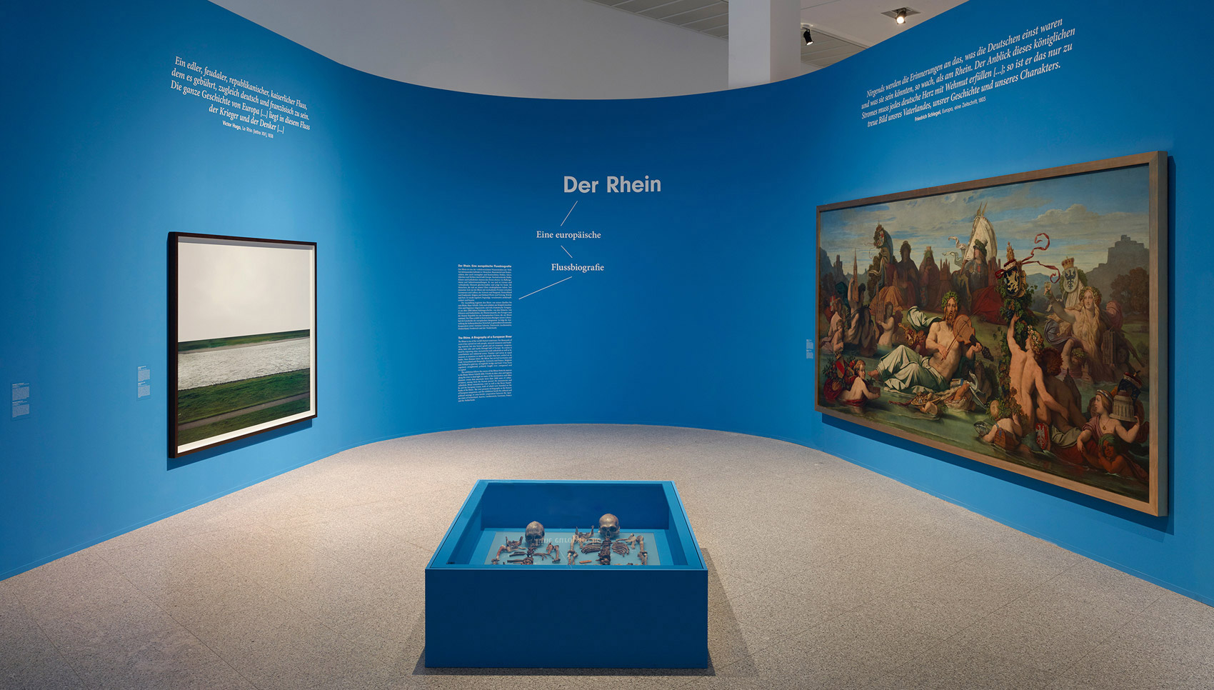 In einem blauen Ausstellungsraum hängen Gemälde. Im Vordergrund steht eine blaue Vitrine mit Exponaten.