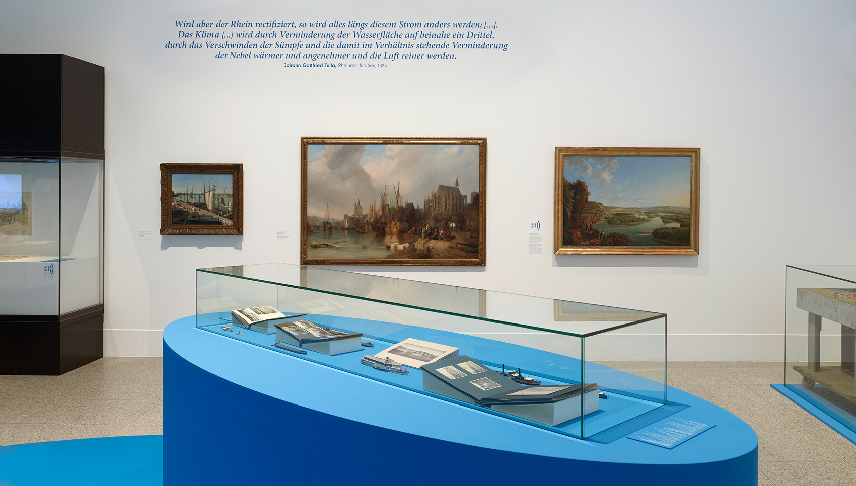 In einem Ausstellungsraum hängen und stehen Exponate. Im Vordergrund steht eine blaue Vitrine mit Exponaten.