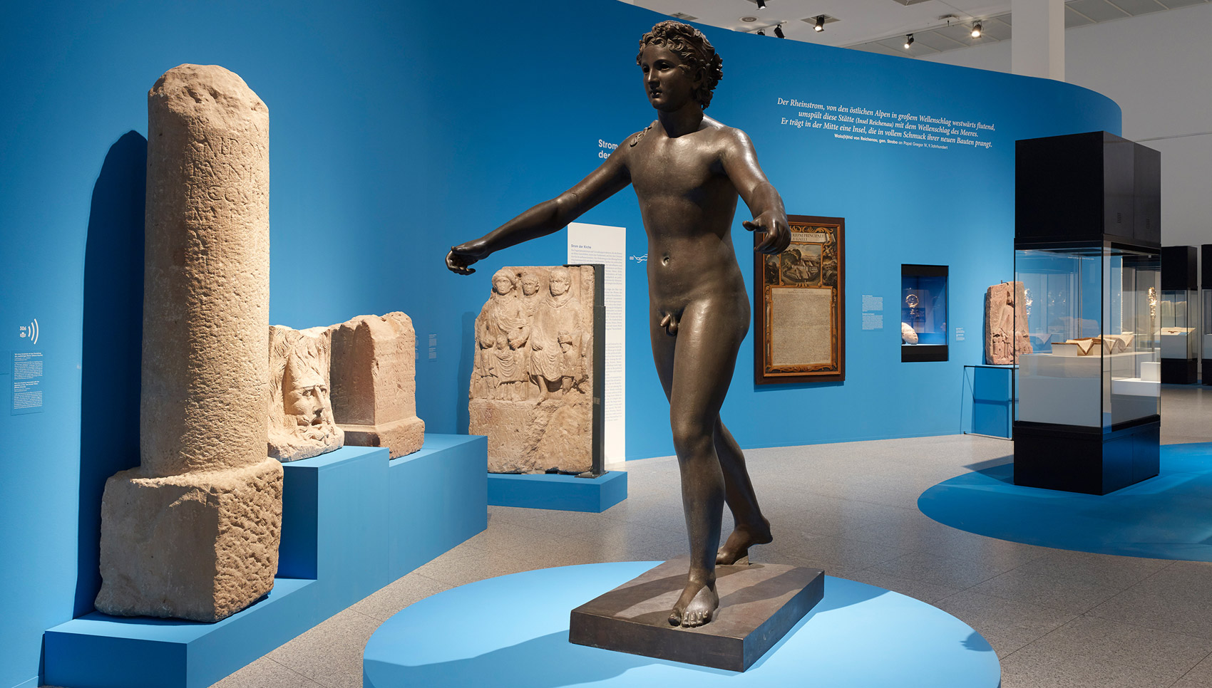 In einem blauen Ausstellungsraum hängen und stehen Exponate. Im Vordergrund steht eine Bronzeskulpur.