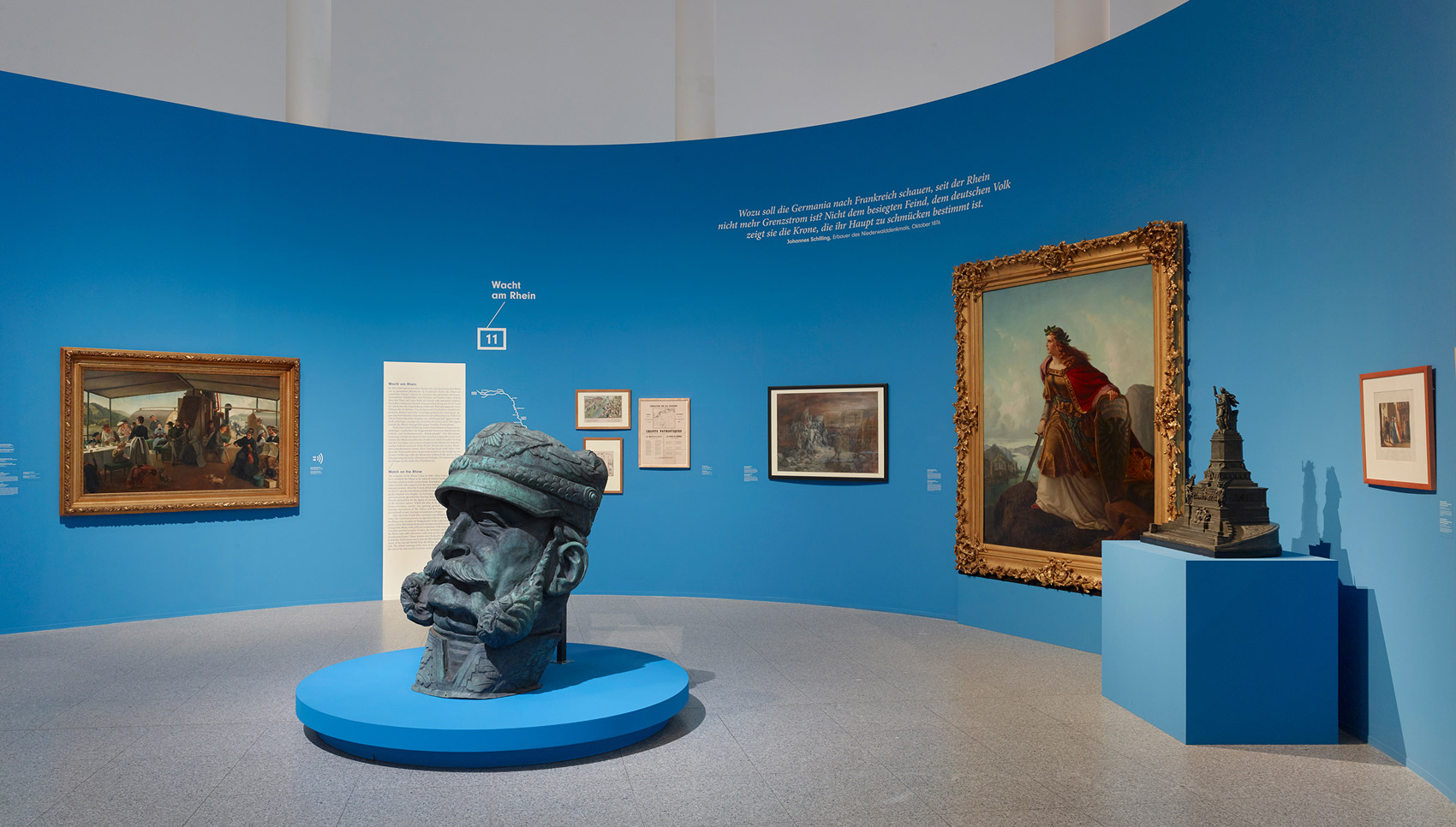 In einem blauen Ausstellungsraum hängen Gemälde und stehen Exponate. In der Mitte steht eine große Kopfskulptur.