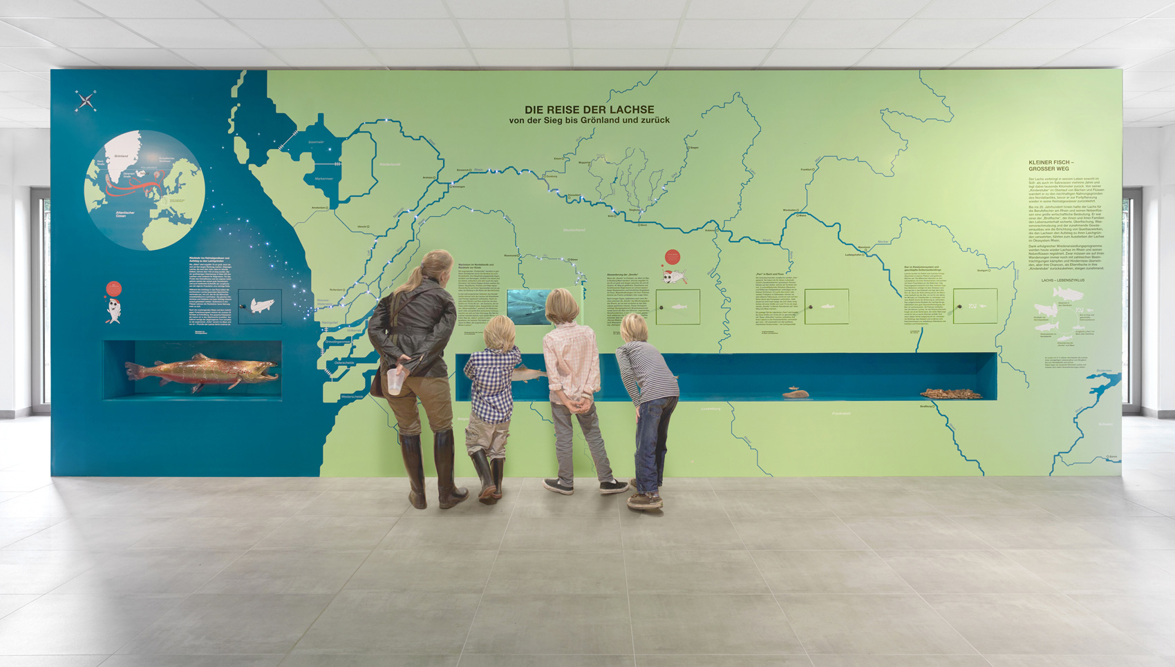 Drei Kinder und eine erwachsene Person stehen im Ausstellungsraum vor einer großen Wandgrafik zum Thema "Die Reise der Lachse"