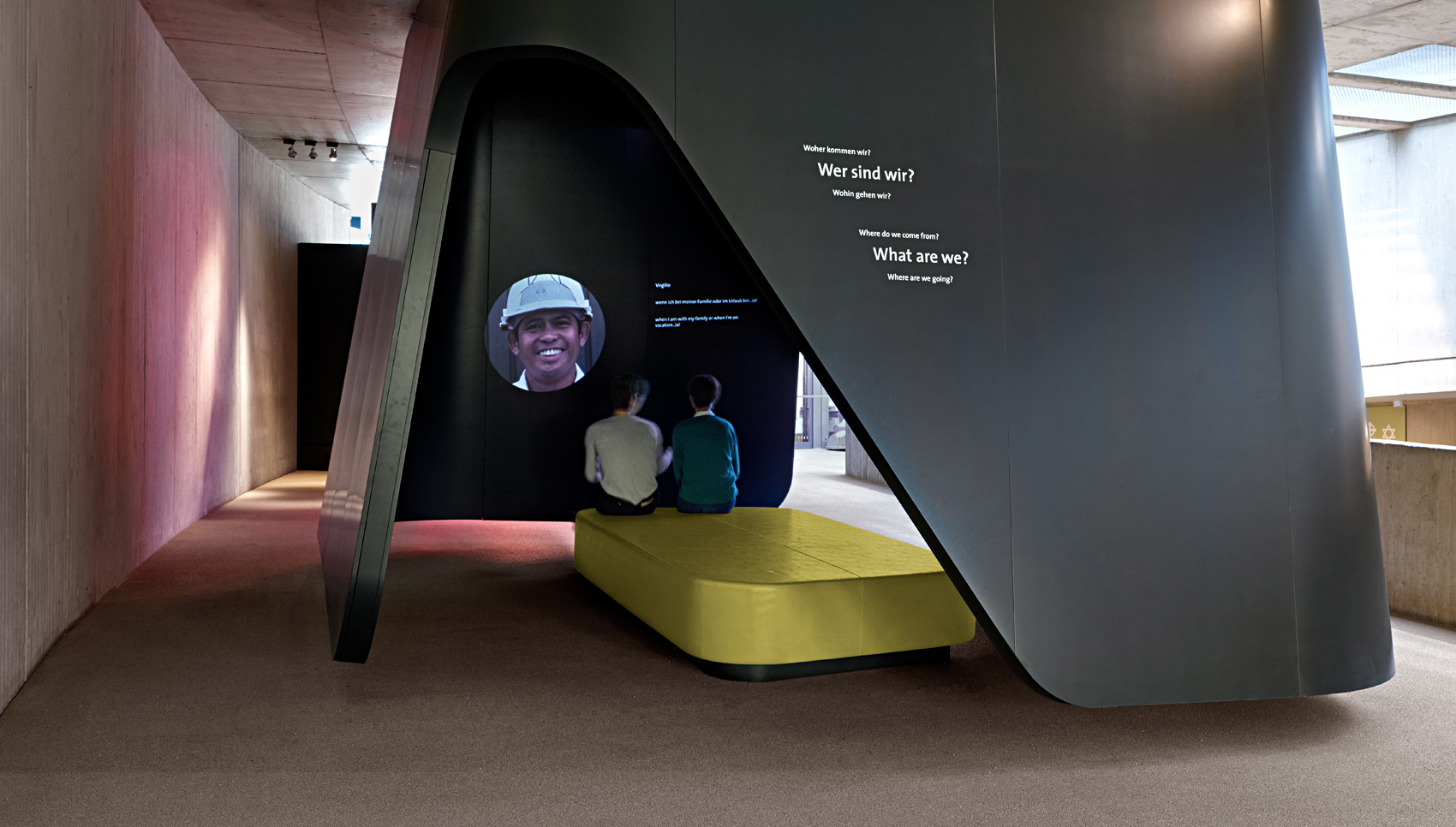 Ausstellungsraum mit gebautem, begehbarem Raum, in dem sich zwei sitzende Menschen eine Projektion anschauen