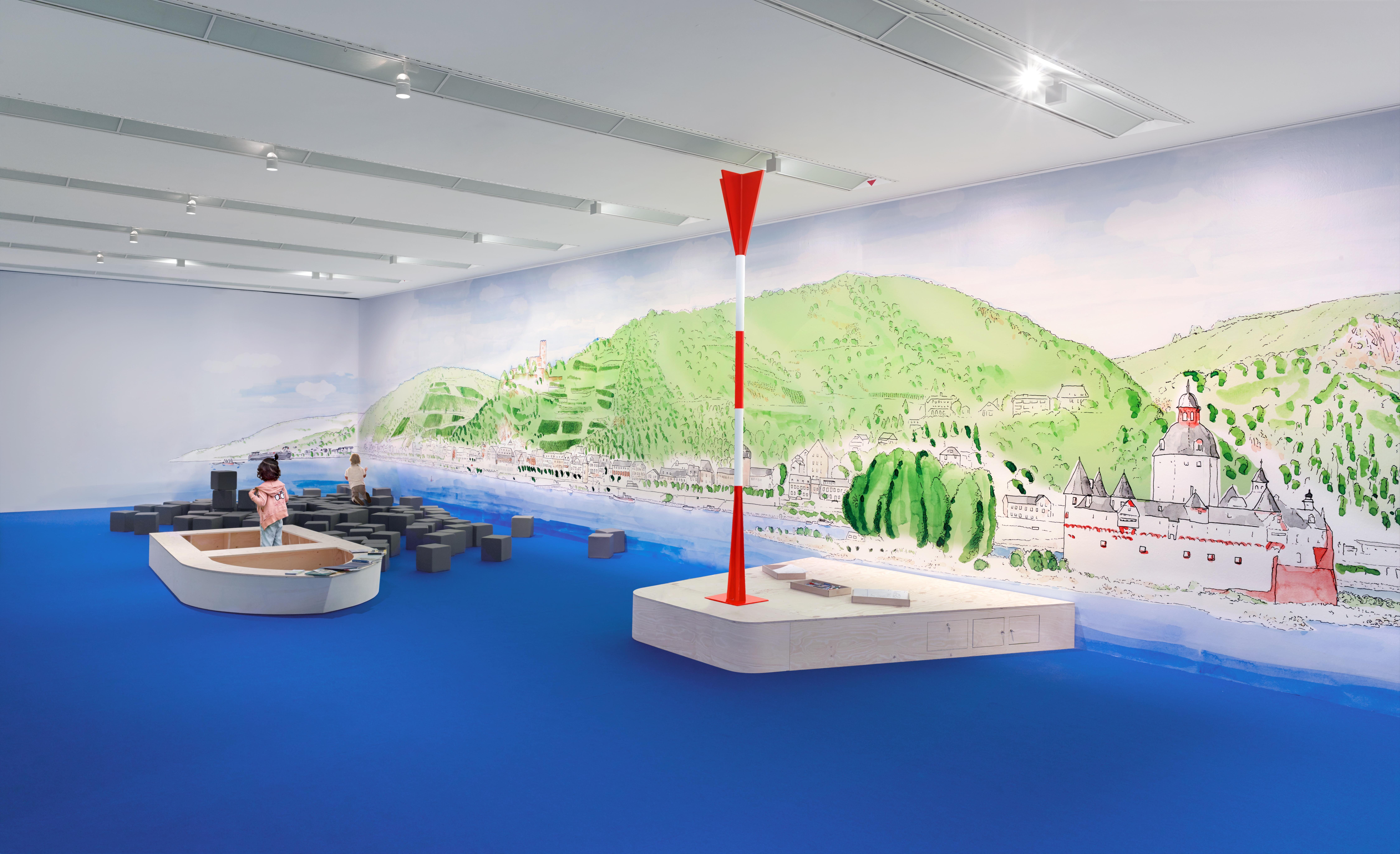 In einem Ausstellungsraum, dessen Boden blau ist, spielen zwei Kinder in nachgebauten Schiffen. Die Wand im Hintergrund hat eine Lanschaftsillustration.