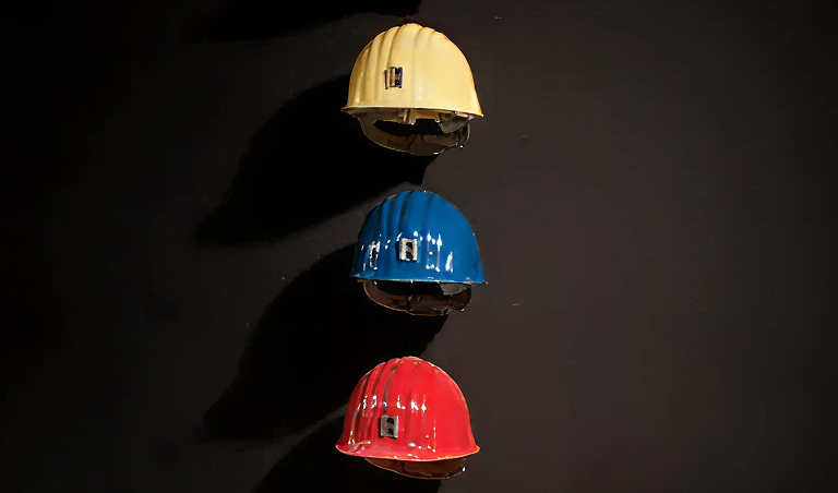 Ausstellungsinstallation, bei der bunte Sicherheutshelme übereinander vor einer schwarzen Wand angeordnet sind