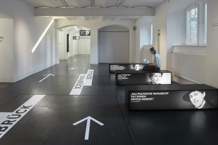 Ausstellungsraum „Stadtspuren Osnabrück“ mit hellen Pfeilen auf dumklem Boden und Sitzgelegenheiten mit Ausstellungsgrafiken