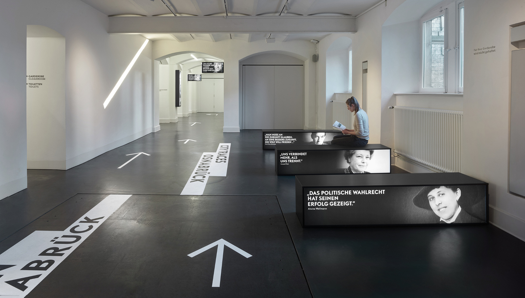 Ausstellungsraum „Stadtspuren Osnabrück“ mit hellen Pfeilen auf dumklem Boden und Sitzgelegenheiten mit Ausstellungsgrafiken