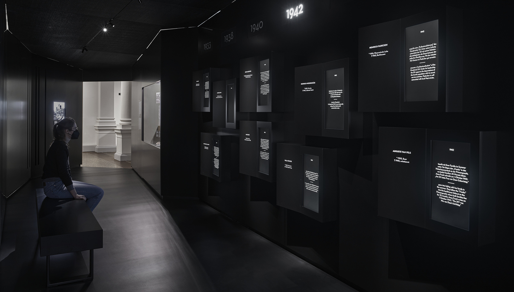 Person sitzt auf Sitgelegenheit und schaut sich mehreren Monitoren auf der gegenüberliegenden schwarzen Wand an