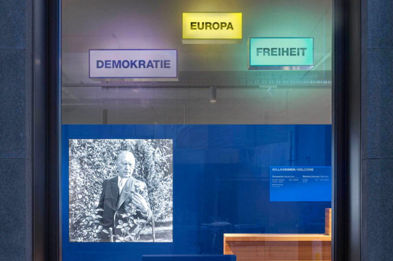 Schaufenster von außen mit 3 Leuchtboxen "Demokratie", "Europa" und "Freiheit". Dahinter eine blaue Wand mit einem großen Display auf dem Konrad-Adenauer in schwarz-weiß zu sehen ist.