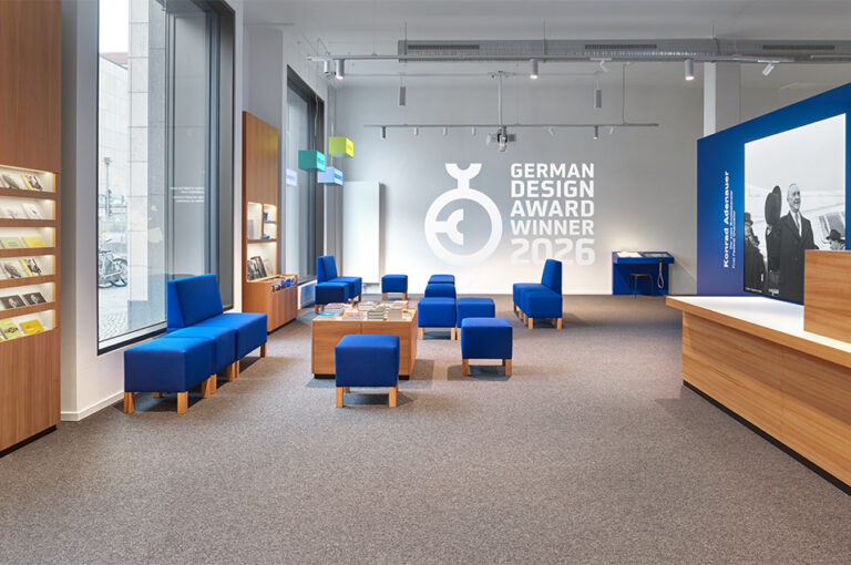 Forum mit rechteckigen, blauen Sitzmöbeln unterschiedlicher Größe. Rechts ein Empfangstresen, dahinter ein großes Bild von Konrad Adenauer. An der zentralen Wand eine Projektion mit Statistiken und einer Europakarte.