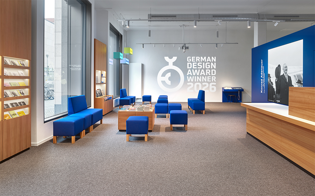 Forum mit rechteckigen, blauen Sitzmöbeln unterschiedlicher Größe. Rechts ein Empfangstresen, dahinter ein großes Bild von Konrad Adenauer. An der zentralen Wand eine Projektion mit Statistiken und einer Europakarte.