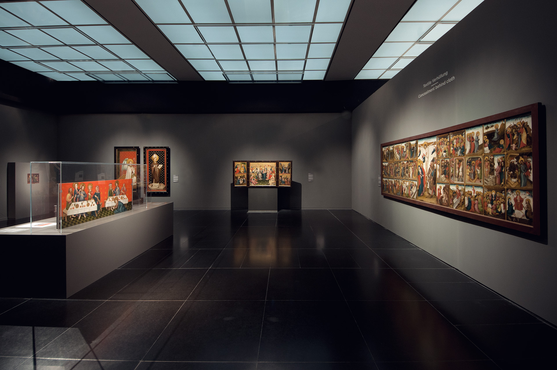 Blick in Ausstellung mit dunkelgrauen Wänden. Links lange Aufsatzvitrine mit Gemälde. Rechts lange Gemälde und Text darüber "Textile Verhüllung"