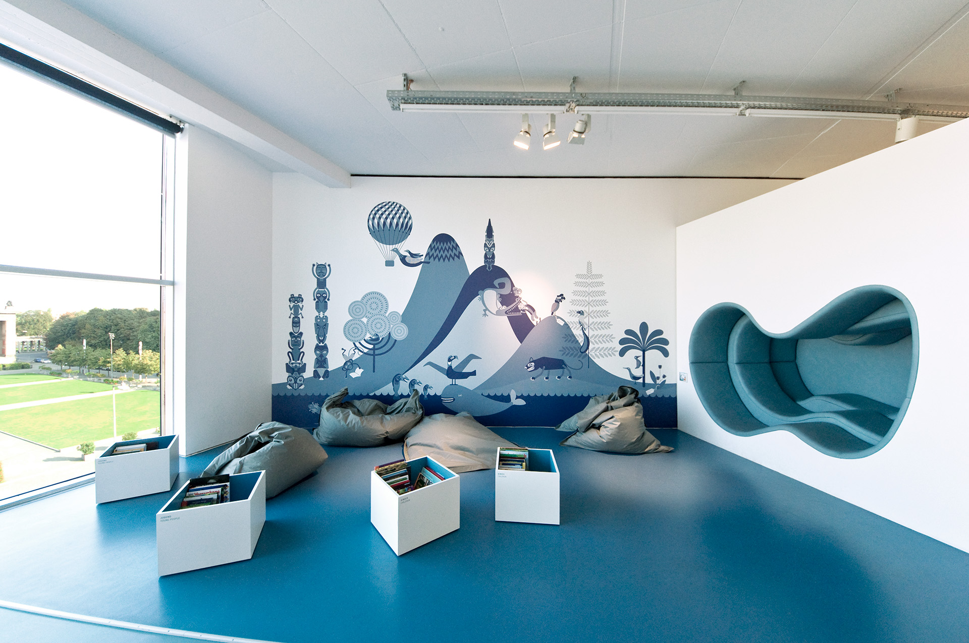 Ausstellungsbereich mit blauem Boden und grauen Sitzkissen. Dazwischen Kisten mit Büchern