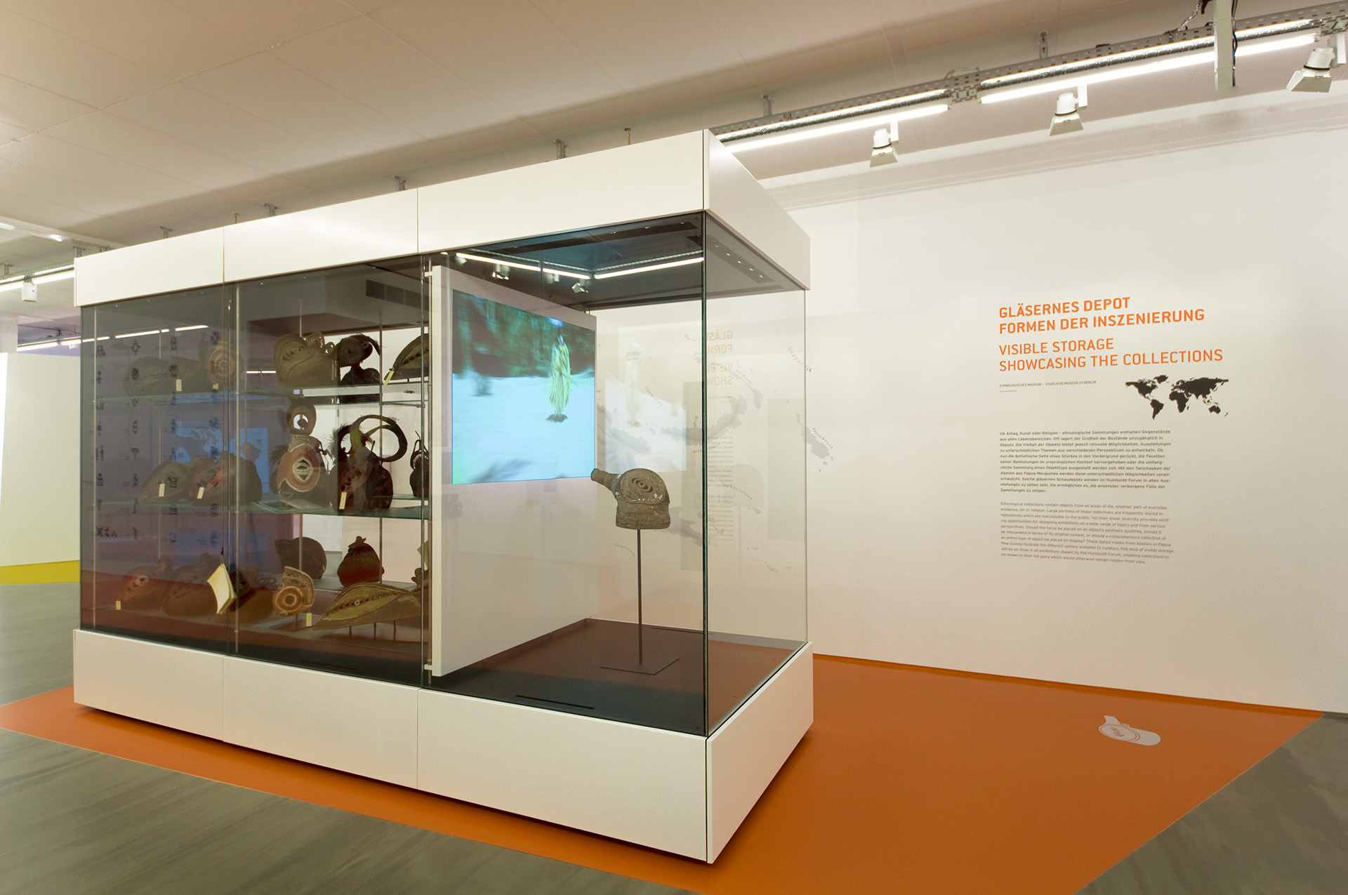 große freistehende Schrankvitrine mit verschiedenen ethnologischen Exponate darin. Wandtext "Gläsernes Depot Formen der Inszenierung"