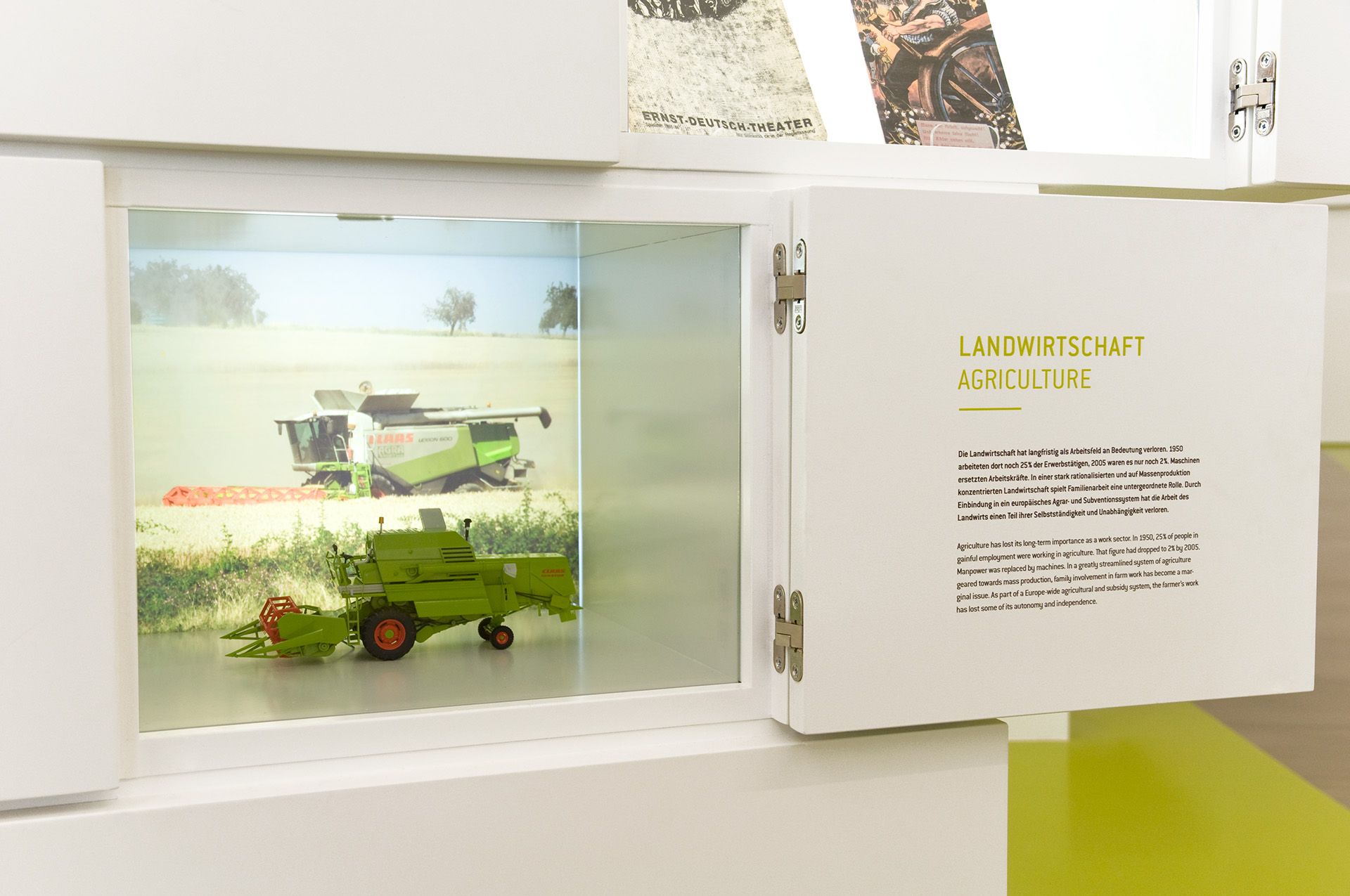 Geöffnete Klappe. Kleines Modell einer Landwirtschaftsmaschine hinter Glas zu sehen. Auf Innenseite der Klappe steht Text zum Thema "Landwirtschaft"