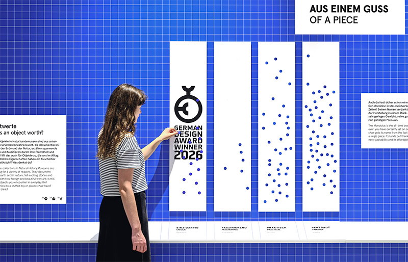Frau, die an einer partizipativen Wand Sticker aufklebt. Über das Foto ist das Logo als German Design Award Winner 2026 gelegt.